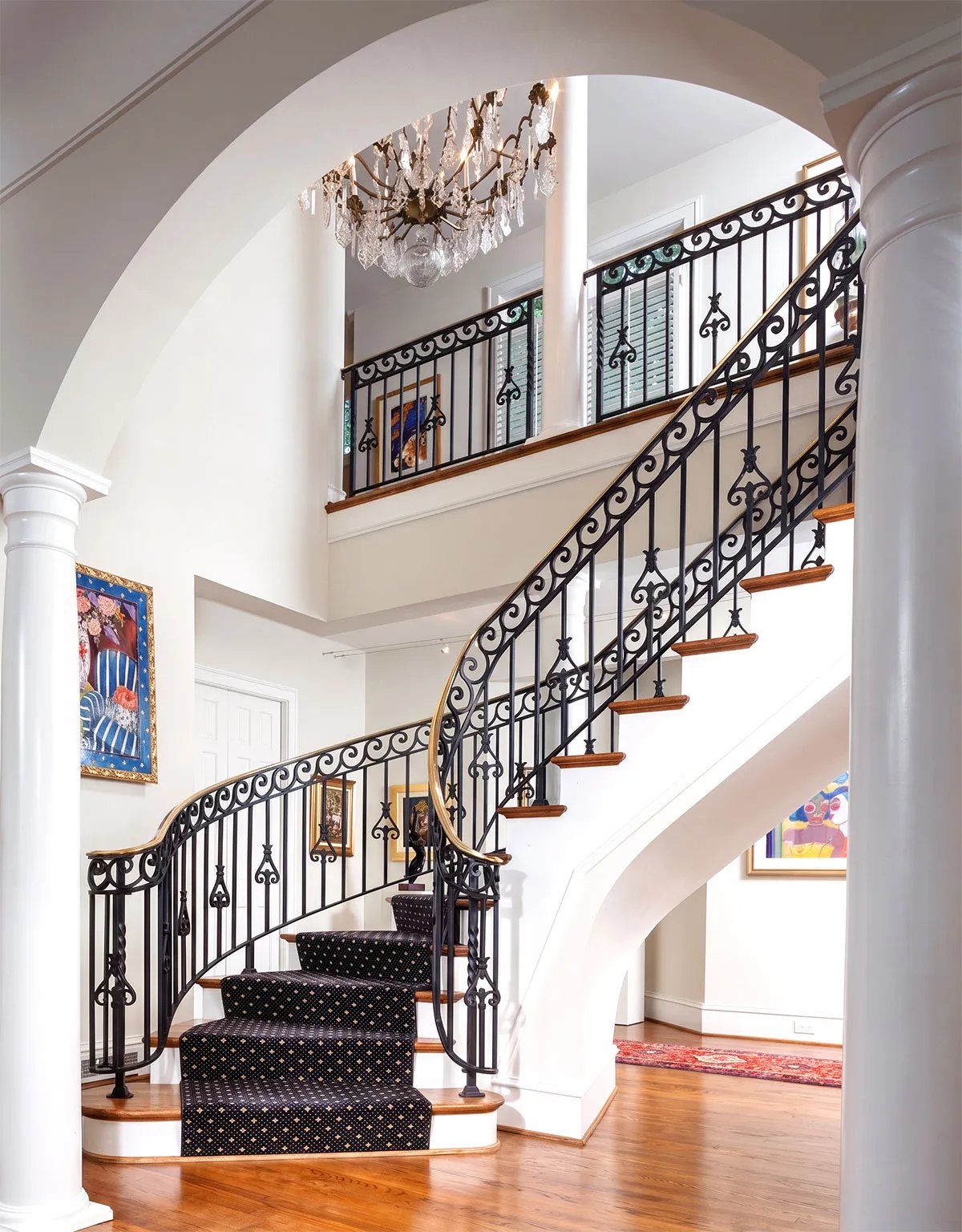 Elegant curved staircase with black wrought iron railing and dark patterned carpet runner, set in a bright foyer with polished wood floors, white walls, white columns, a large chandelier, and colorful framed artwork.