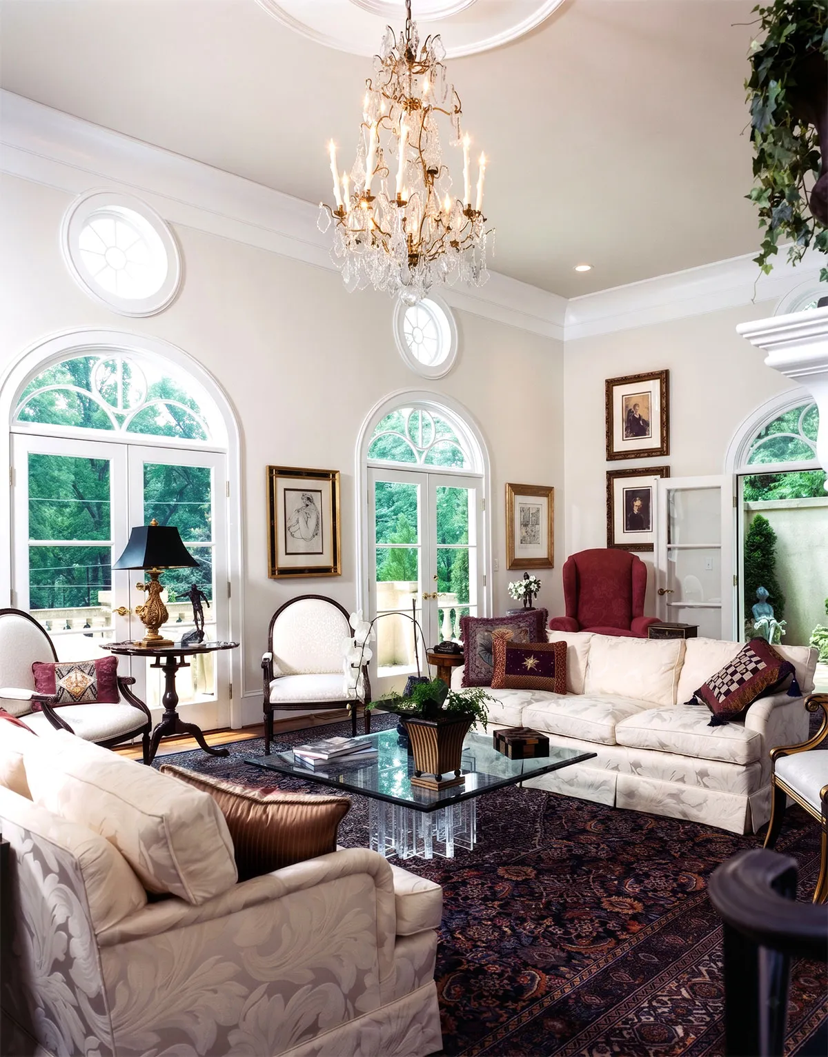 Elegant living room with white upholstered sofas and chairs, a glass coffee table with plants and books, a decorative dark patterned rug, and a crystal chandelier hanging from the ceiling. Large arched windows and French doors let in natural light, showing green trees outside. Wall art and framed pictures adorn the walls, and a red armchair is in the corner near an open door leading to an outdoor patio with greenery.