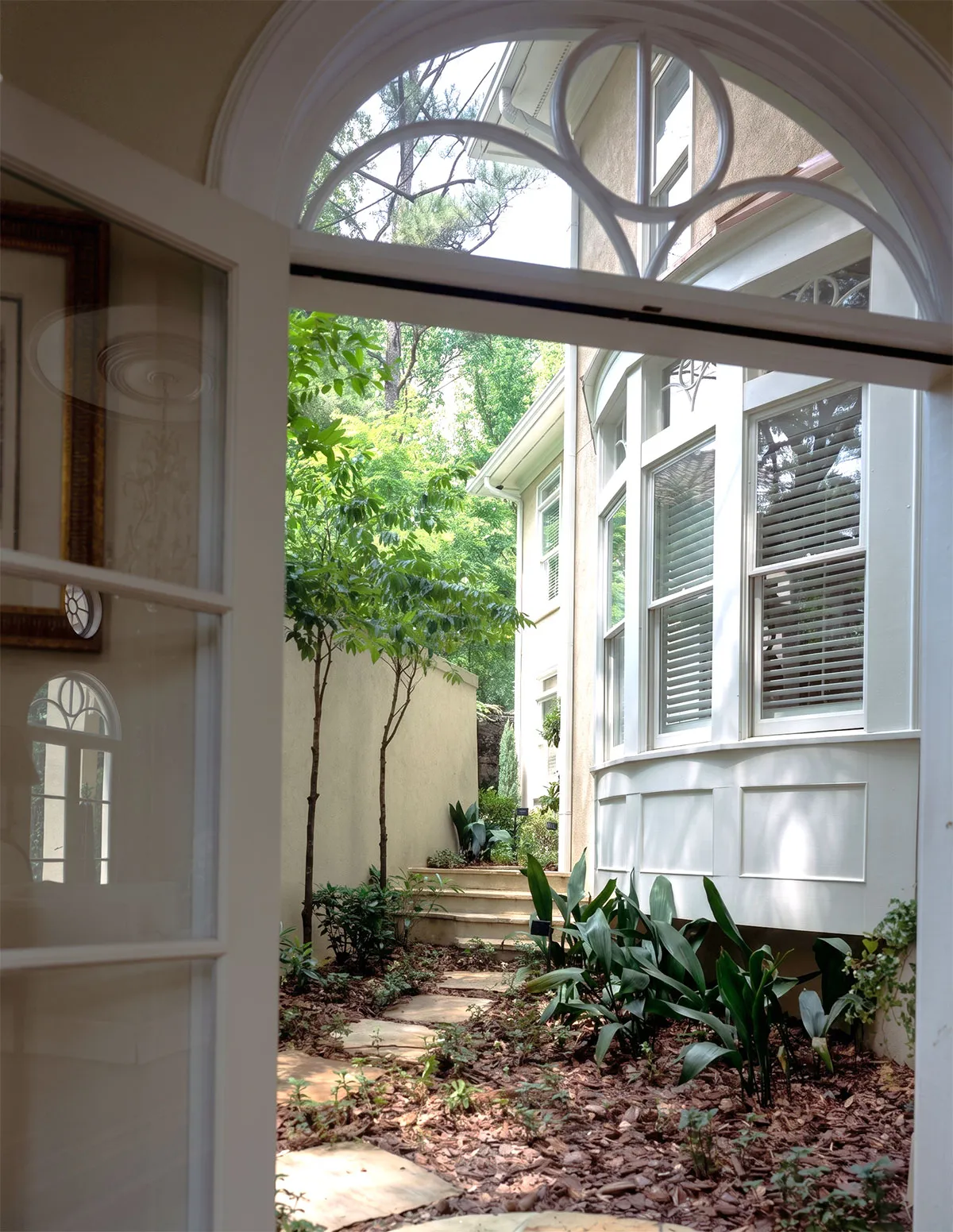 View through an open white door showing a stone pathway bordered by plants and small trees leading to steps beside a house with white framed windows and beige walls.