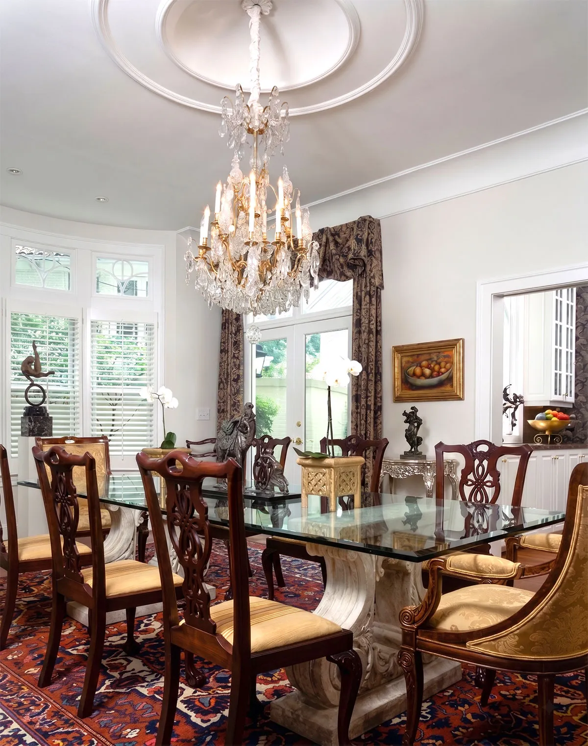 Elegant dining room with a large glass-top table supported by ornate stone pedestals, surrounded by wooden chairs with carved backs and upholstered seats, a gold-toned armchair on one end, a crystal chandelier hanging from a decorated ceiling medallion, patterned area rug, sculptures and plants on the table, large windows with white shutters, dark patterned curtains framing glass doors, a fruit painting on the wall, and a view into a kitchen with fruit bowl and cabinetry.