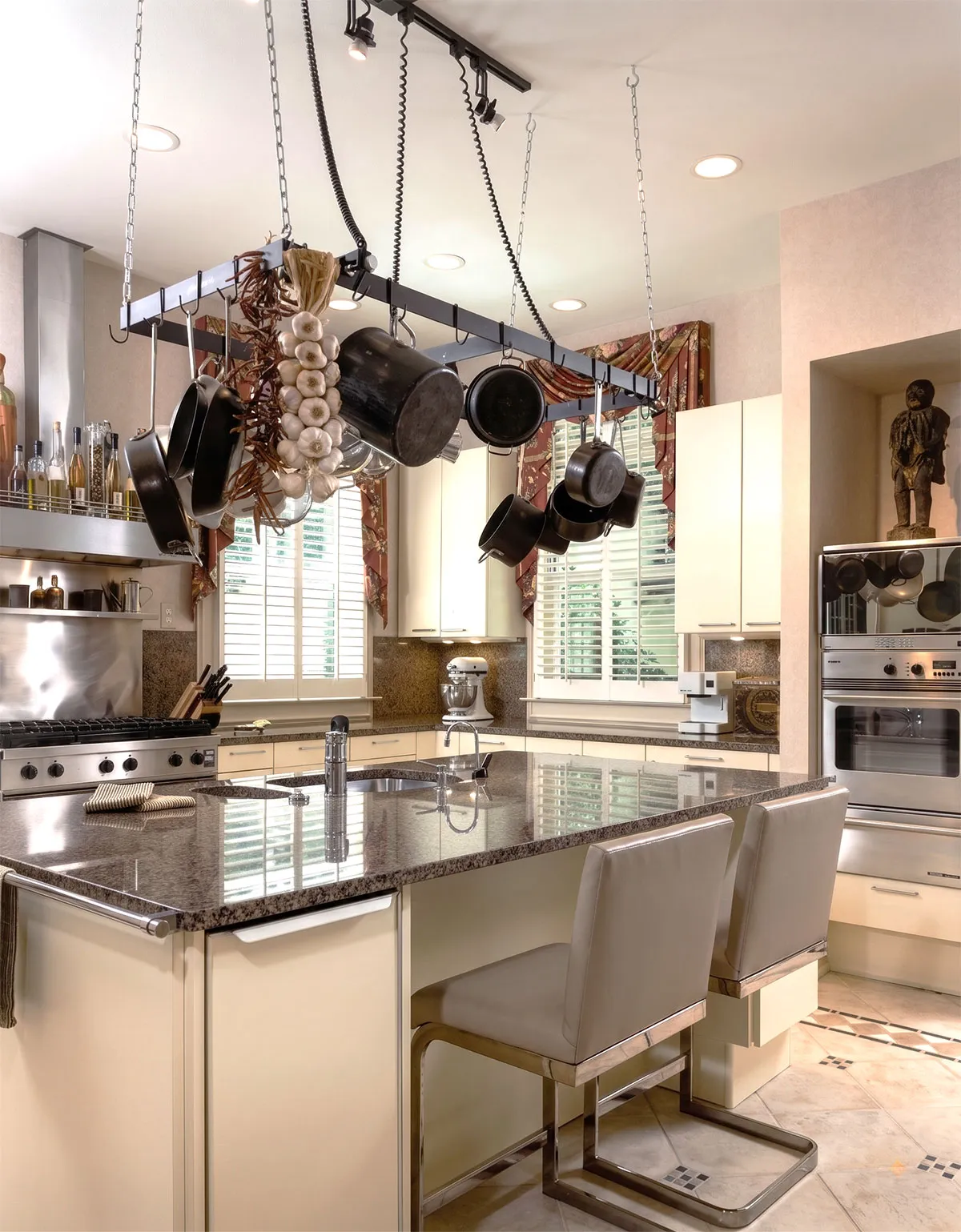 Bright modern kitchen with a large granite island countertop featuring a built-in sink and two gray cushioned bar stools, hanging pot rack with various pots, pans, garlic, and dried peppers above the island, stainless steel stove and oven, white cabinets, two windows with white shutters and floral valances, a mixer on the counter, and tiled floor with decorative insets.