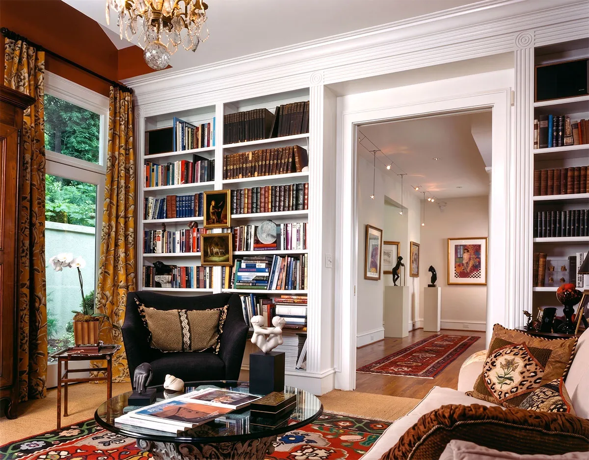 Cozy living room with a black armchair and beige sofa adorned with patterned pillows, a round glass coffee table with books and sculptures, white built-in bookshelves filled with books and framed art, vibrant yellow floral curtains, a wooden side table with a white orchid plant, and a view into a hallway decorated with framed artwork and sculptures on pedestals, with wooden floors and an oriental runner rug.