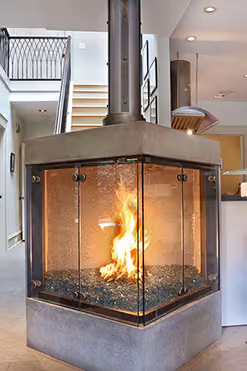 Modern corner fireplace with glass panels and a fire burning over blue decorative stones in a bright home interior.