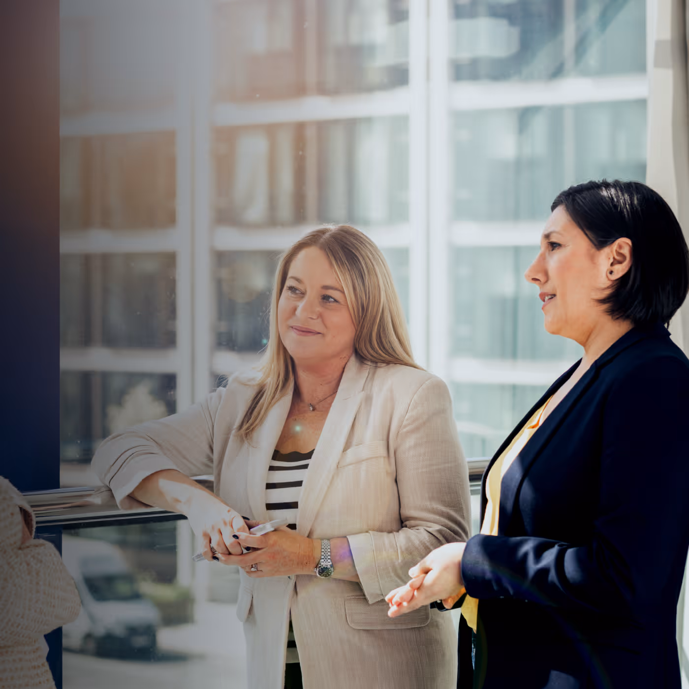Two women in business attire having a friendly conversation by a window in a modern office.