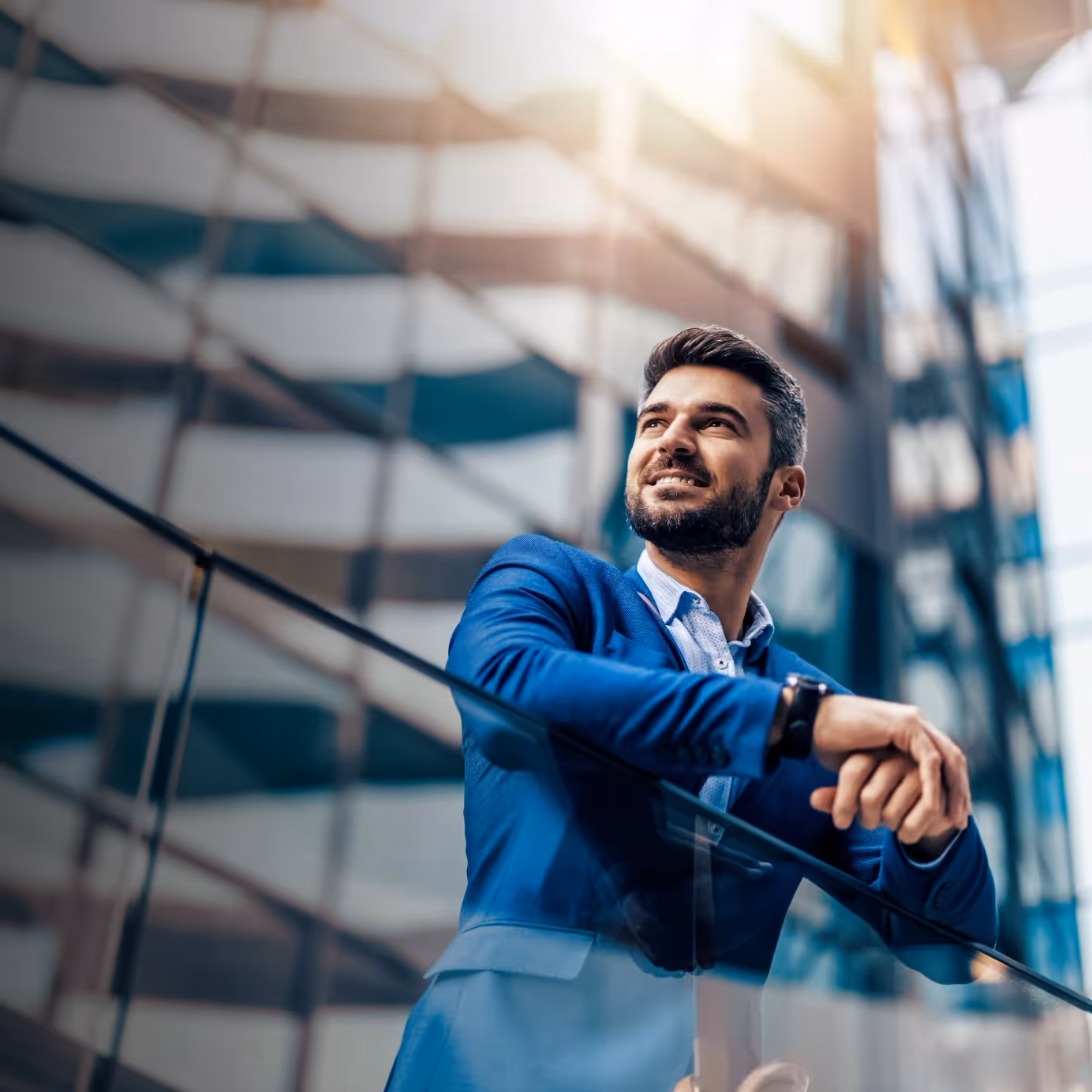 Smiling man in a blue suit leaning on a glass railing in front of a modern building.
