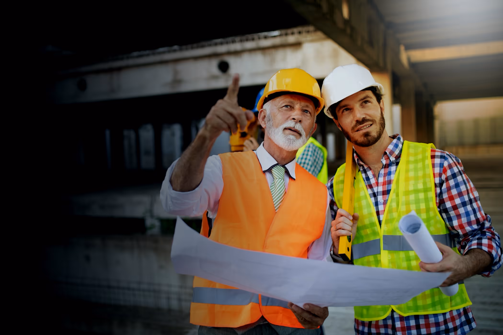 Two construction workers in helmets and vests examine blueprints at a worksite under a structure.