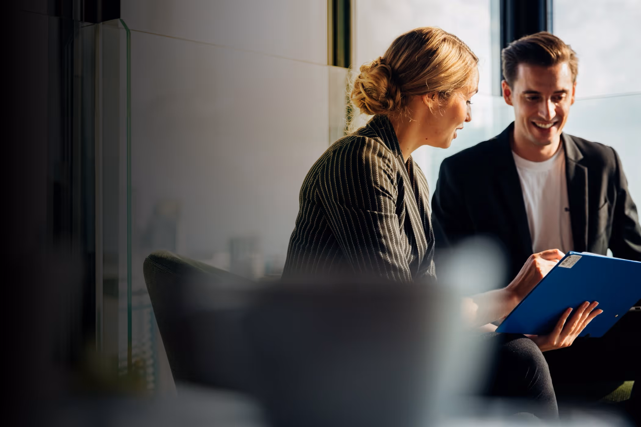 Two business people smiling and reviewing documents on a blue clipboard in an office.
