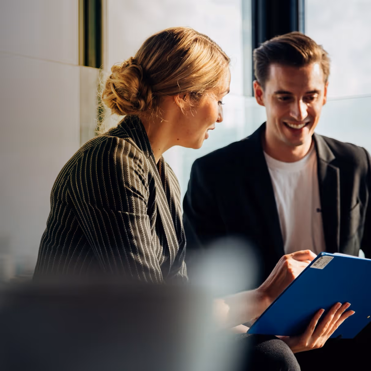 Smiling man and woman reviewing documents on a blue clipboard in a modern office with large windows.