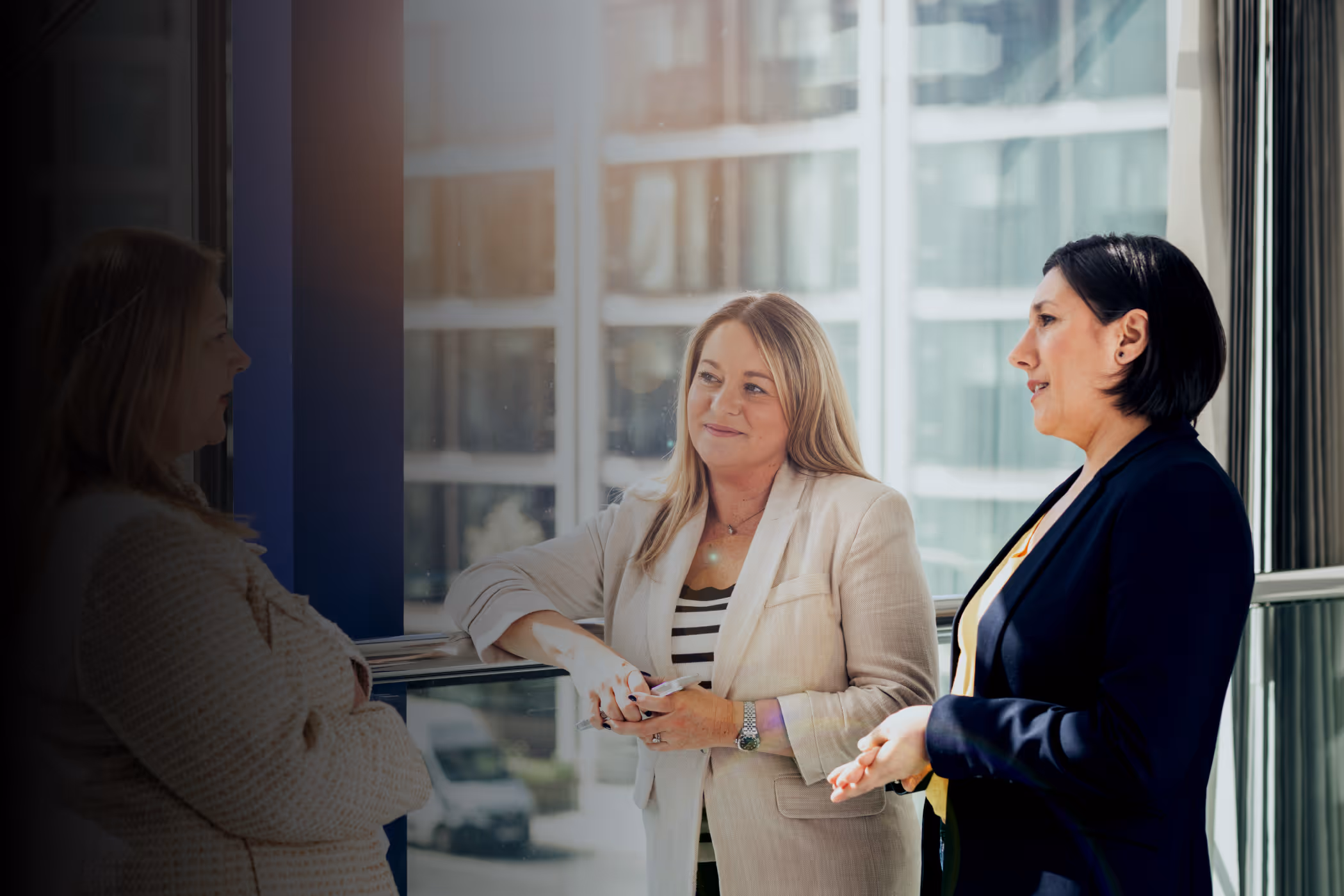 Three professional women in business attire talking in a modern office hallway next to glass windows.