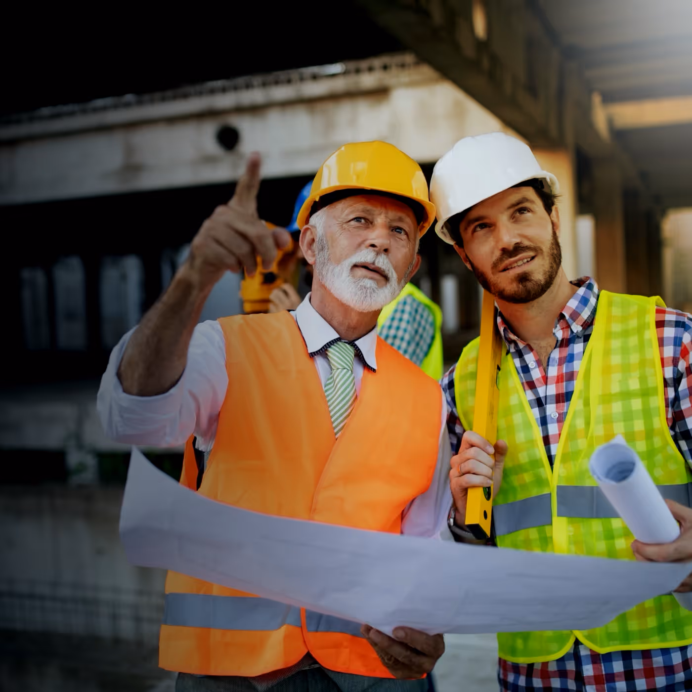 Two construction workers wearing hard hats and reflective vests reviewing blueprints at a site.