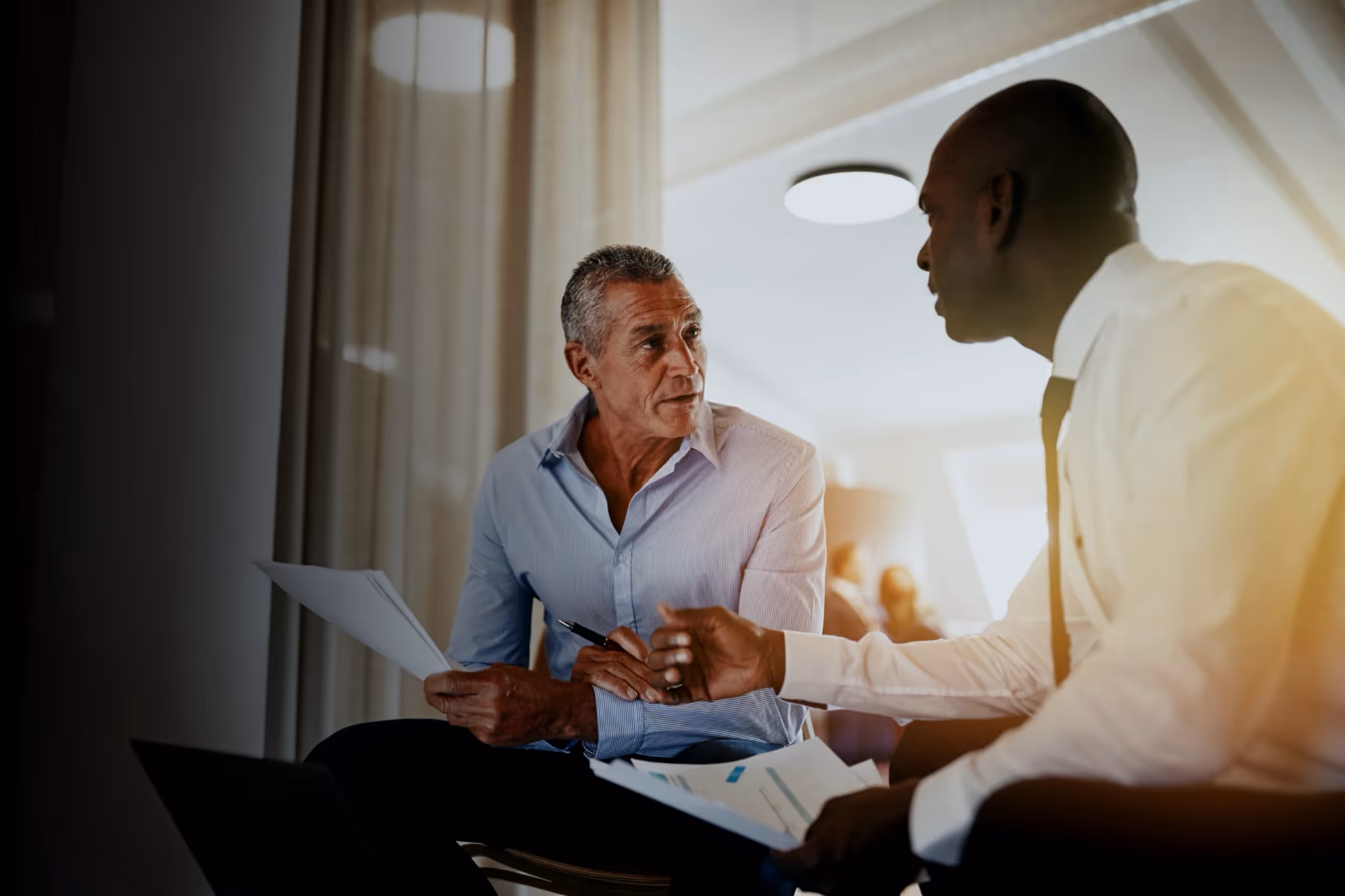 Two men in business attire discussing documents in a bright office setting.