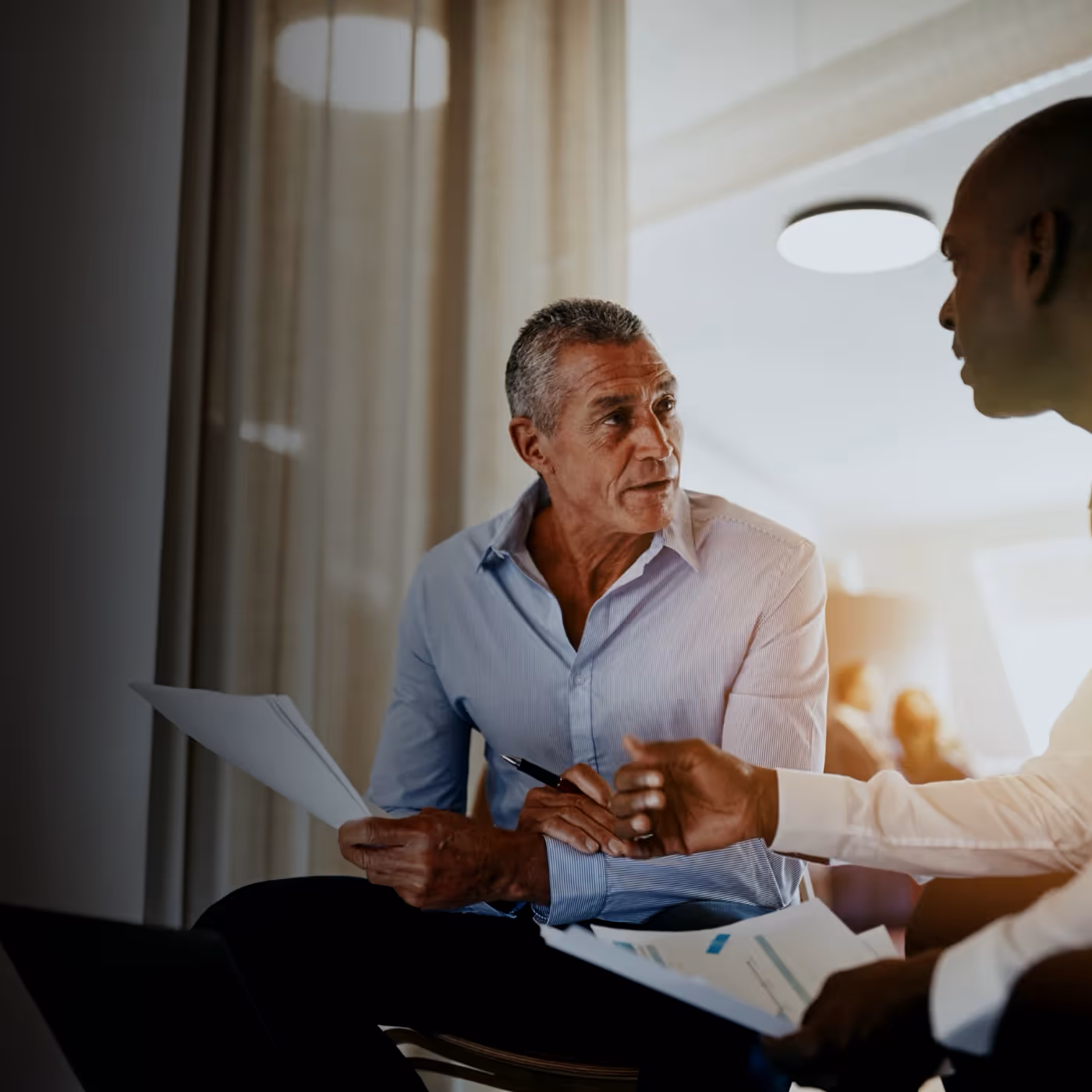 Two men having a discussion while holding documents and a pen in an office setting.