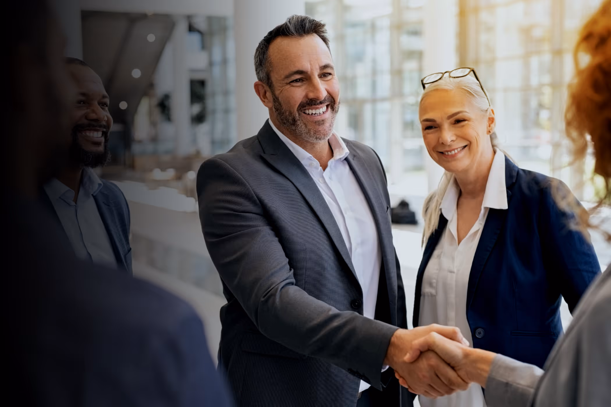 Smiling businesspeople shaking hands in a bright modern office while two colleagues look on.