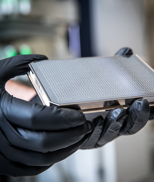 Person wearing black gloves holding a metallic electronic device with a grid of small perforations.