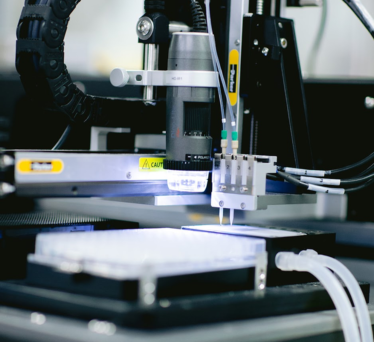 Close-up of a robotic lab machine with precision tubing and a lighted camera focusing on a platform with tubes attached.