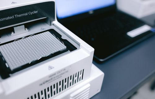 Automated thermal cycler machine with a 96-well PCR plate inside and a laptop in the background.