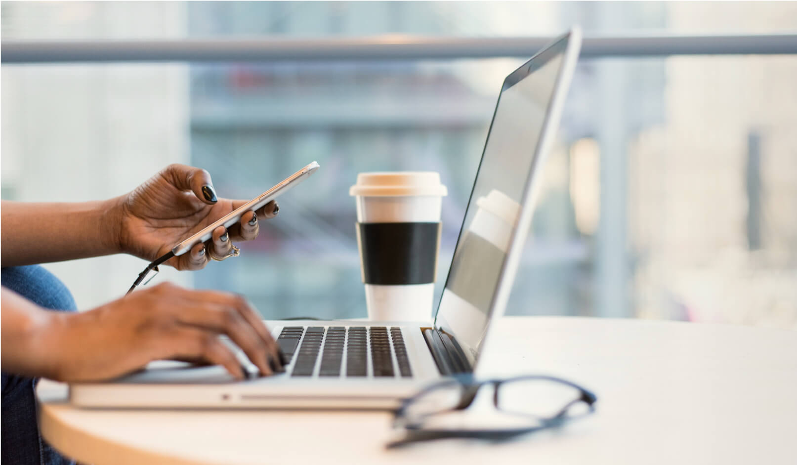 Person using a smartphone and typing on a laptop with a coffee cup and eyeglasses on a white table.