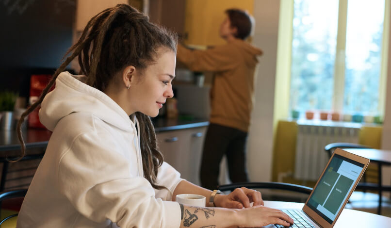 Young woman with dreadlocks and a white hoodie working on a laptop at a table with a coffee cup, while another person in a brown hoodie reaches for a shelf in the background.