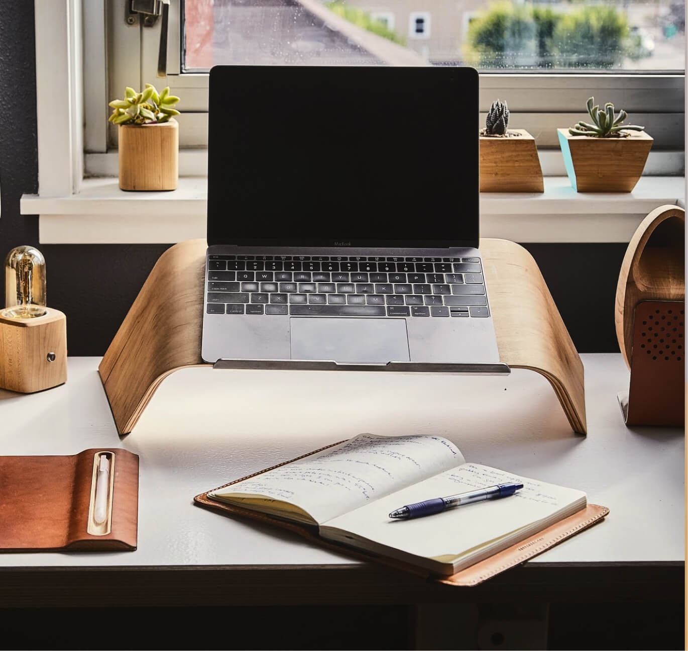Desk setup with a MacBook on a wooden stand, an open notebook with a pen, small potted succulents by a window, and a wooden lamp.