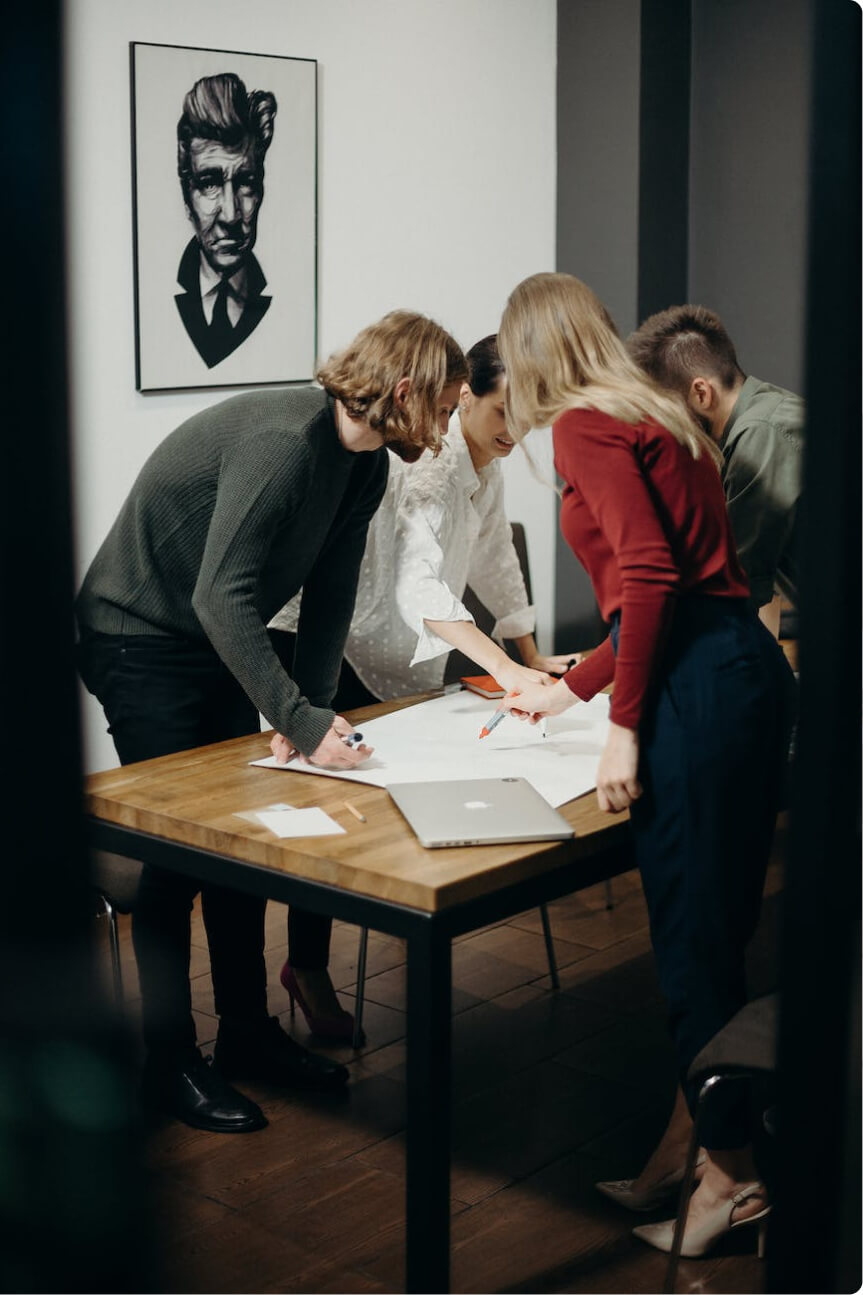 Four young professionals collaborating over a large sheet of paper on a wooden table in a modern office.