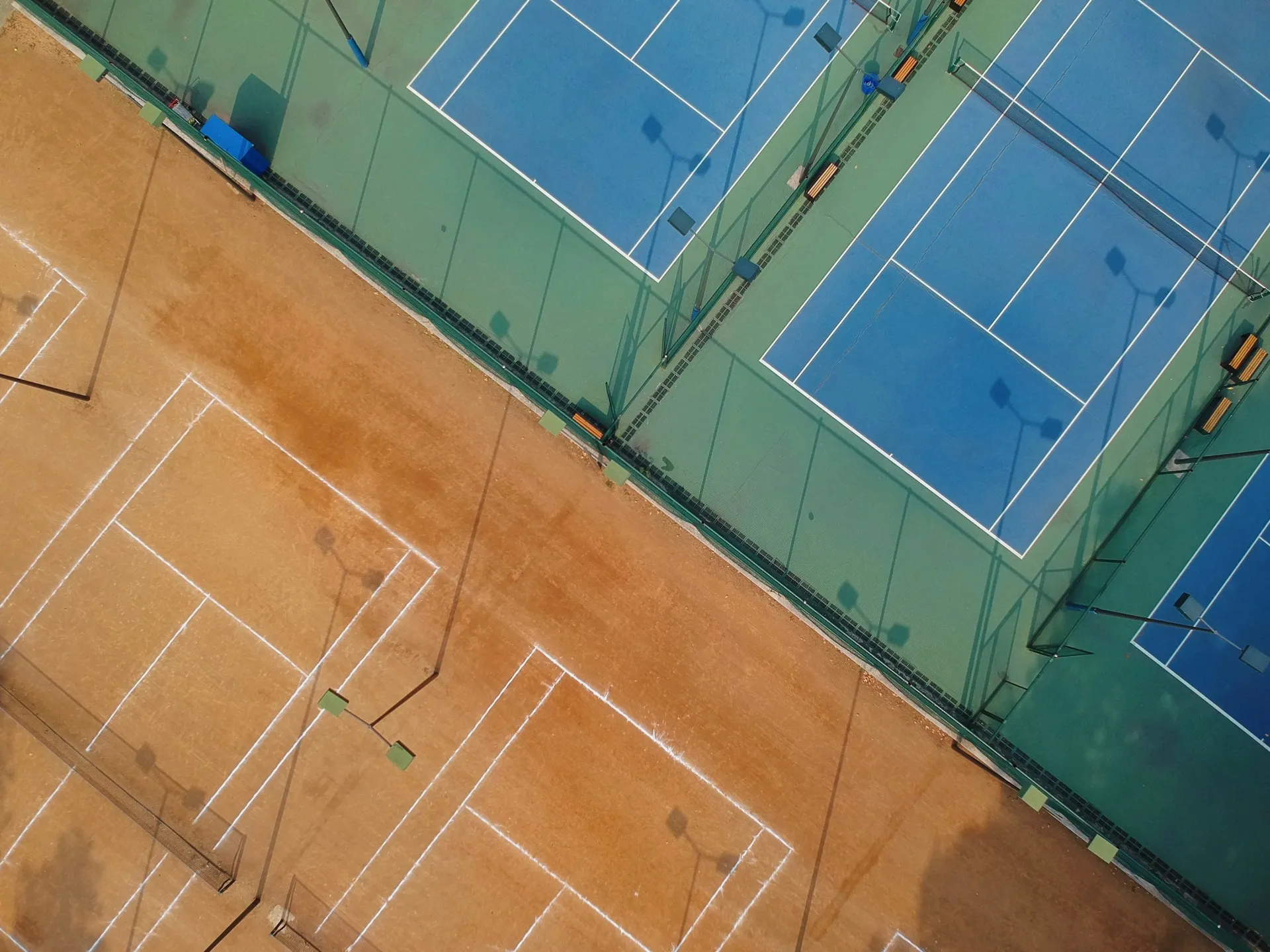 Aerial view of adjacent tennis courts with clay courts on the left and blue hard courts on the right, surrounded by green borders and fencing.