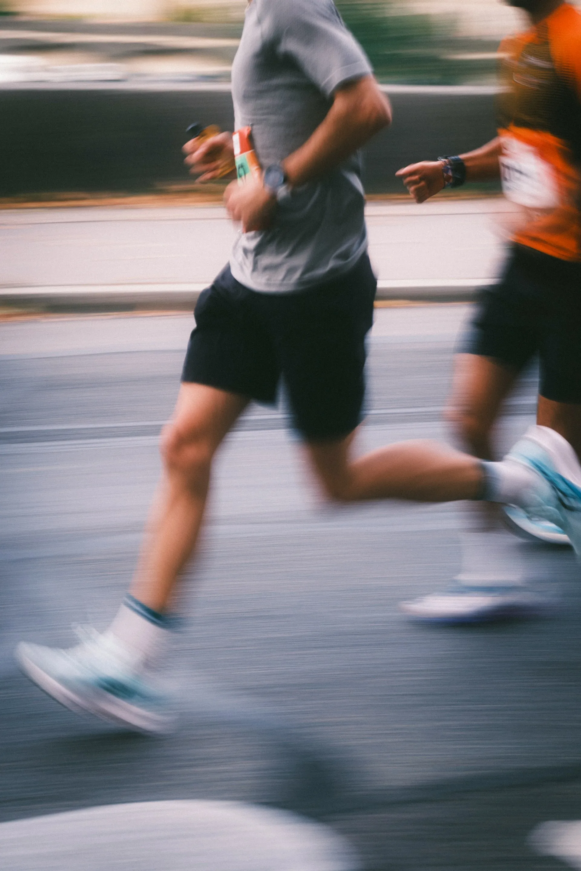Two runners in motion on a road, one wearing a gray shirt and black shorts, the other in an orange shirt.