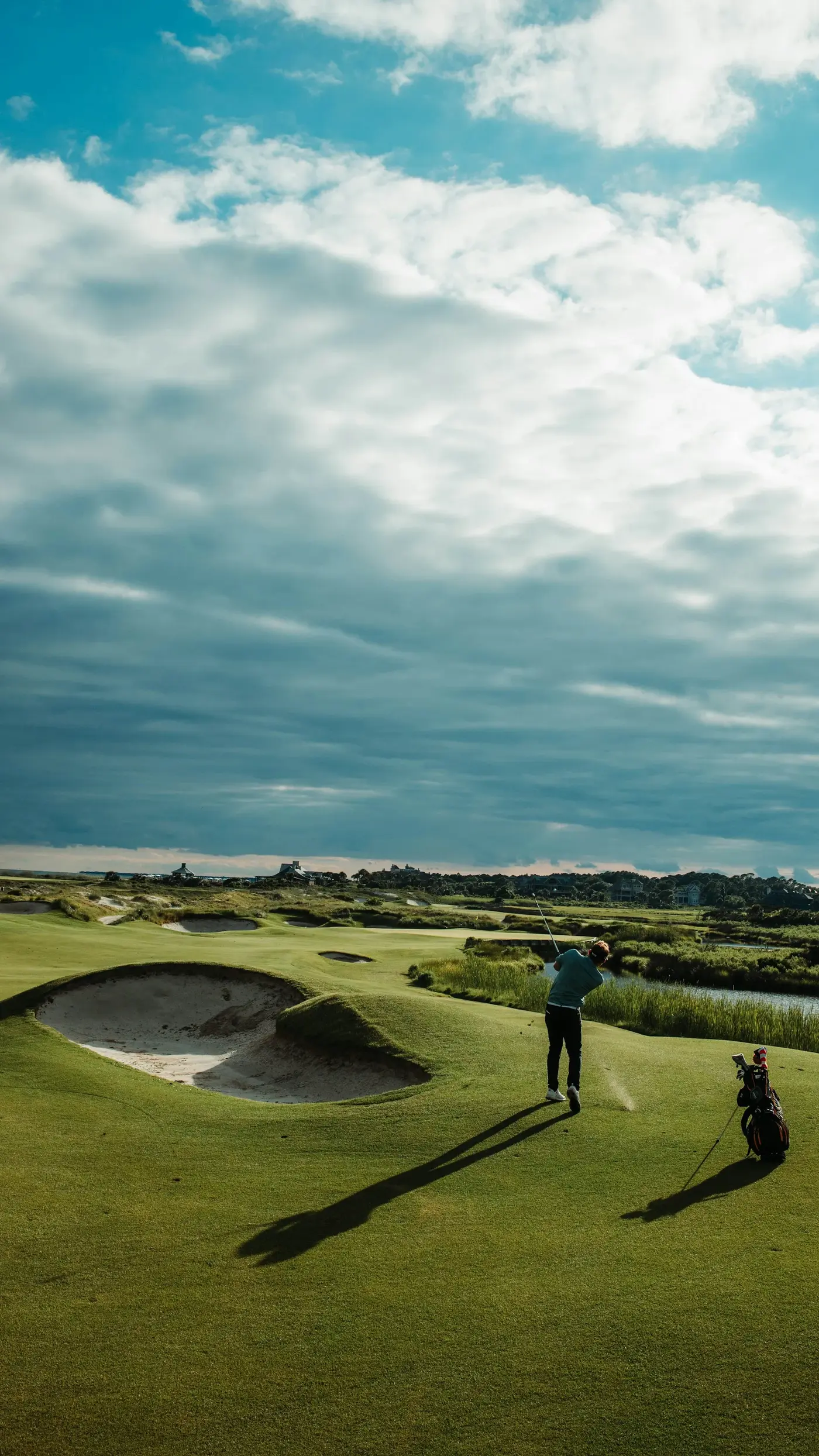 Golfer swinging on a green golf course next to a sand bunker under a partly cloudy sky.