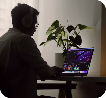 Personne portant un casque et regardant un ordinateur portable avec des logiciels de montage vidéo, avec une plante verte en arrière-plan.