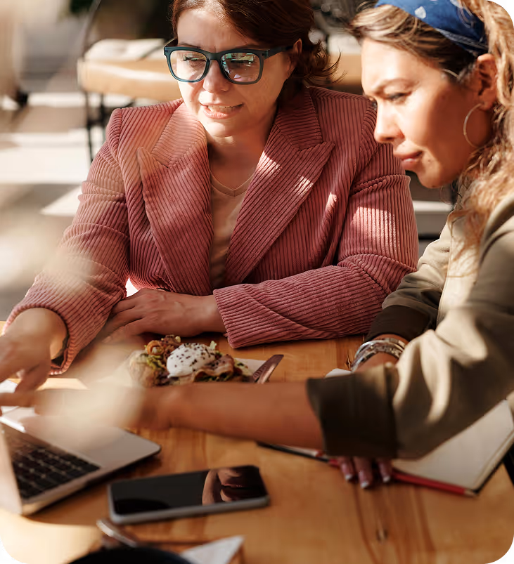 Deux femmes regardant un ordinateur portable sur une table en bois avec un smartphone, un carnet et une assiette de nourriture.