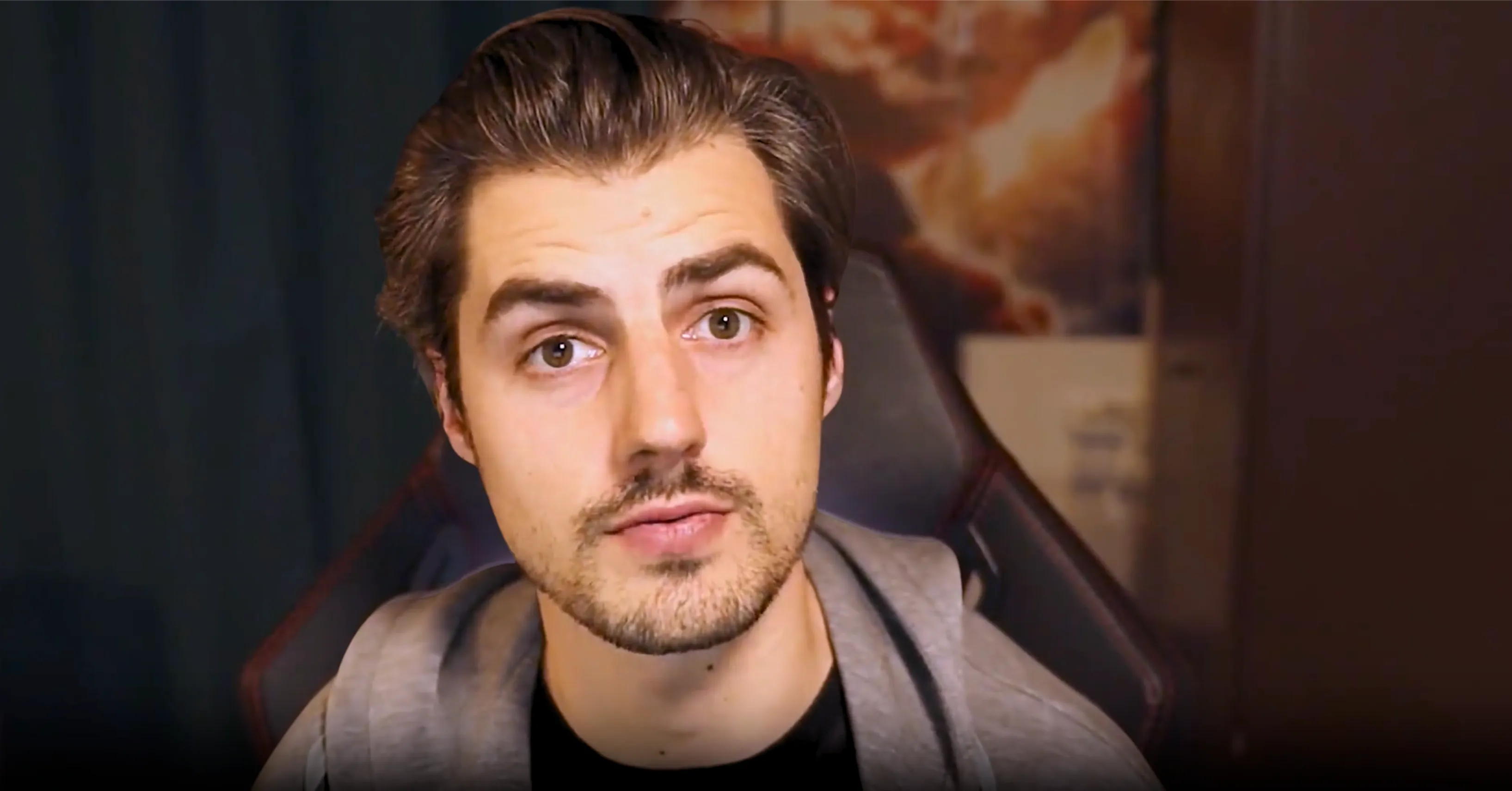 Jokariz - Homme à cheveux bruns courts et barbe légère portant un sweat gris, assis dans un fauteuil gaming noir avec un arrière-plan flou.