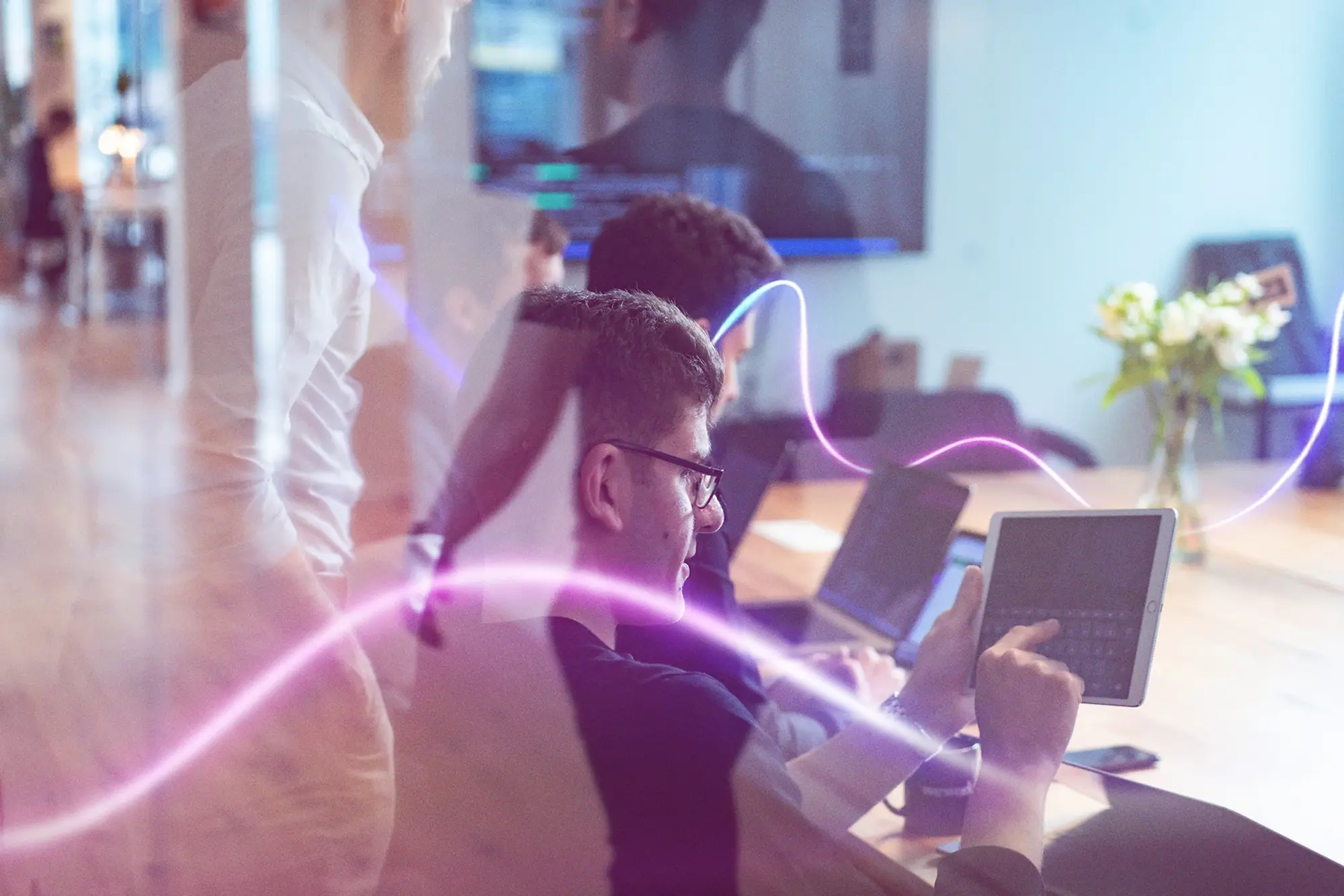 Man using a tablet with others working on laptops around a conference table in a modern office.