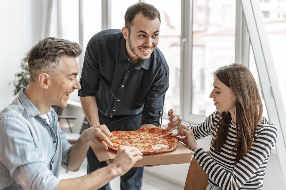 Drei Geschäftspersonen in einem modernen Büro, die gemeinsam eine Pizza aus einer Pizzaschachtel nehmen.