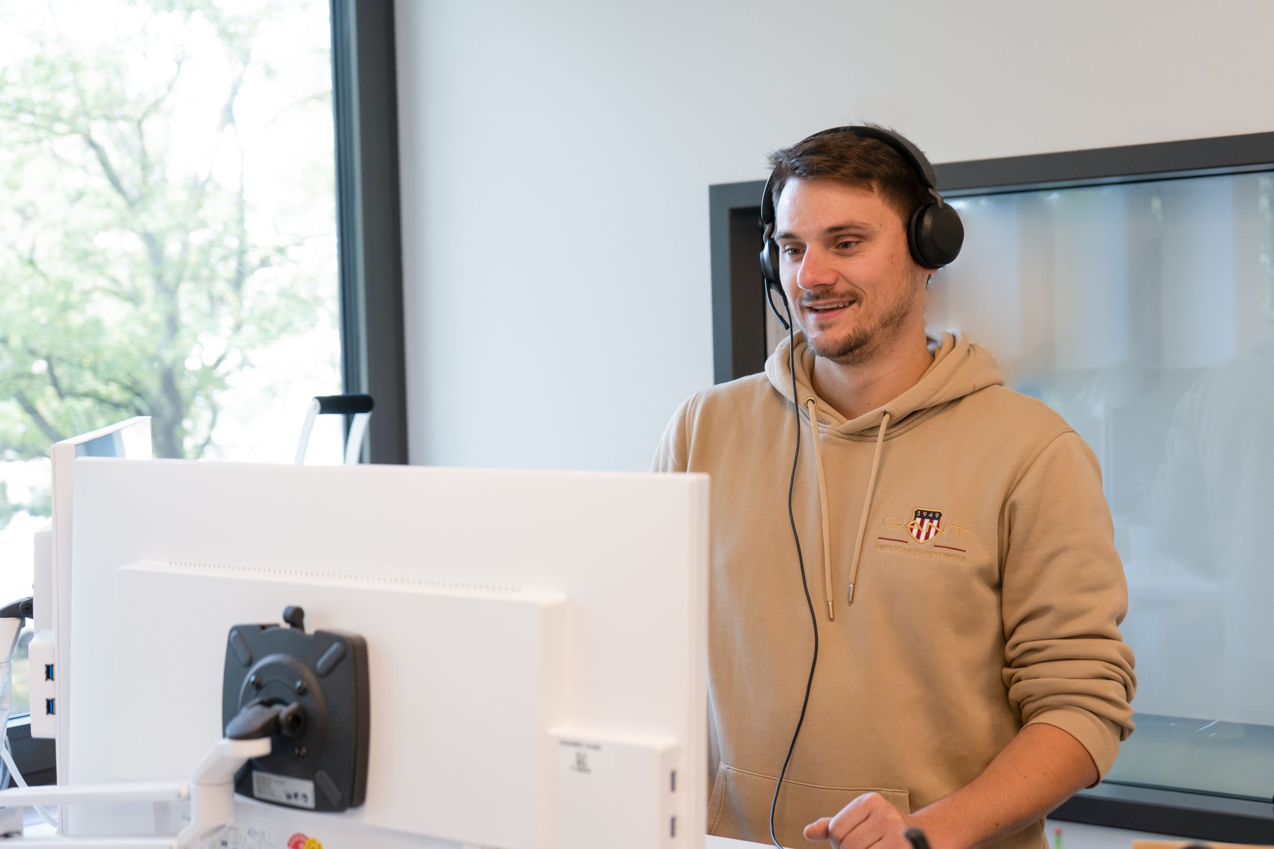 Ein junger Mann mit Headset steht an einem Schreibtisch und schaut auf einen Computerbildschirm.