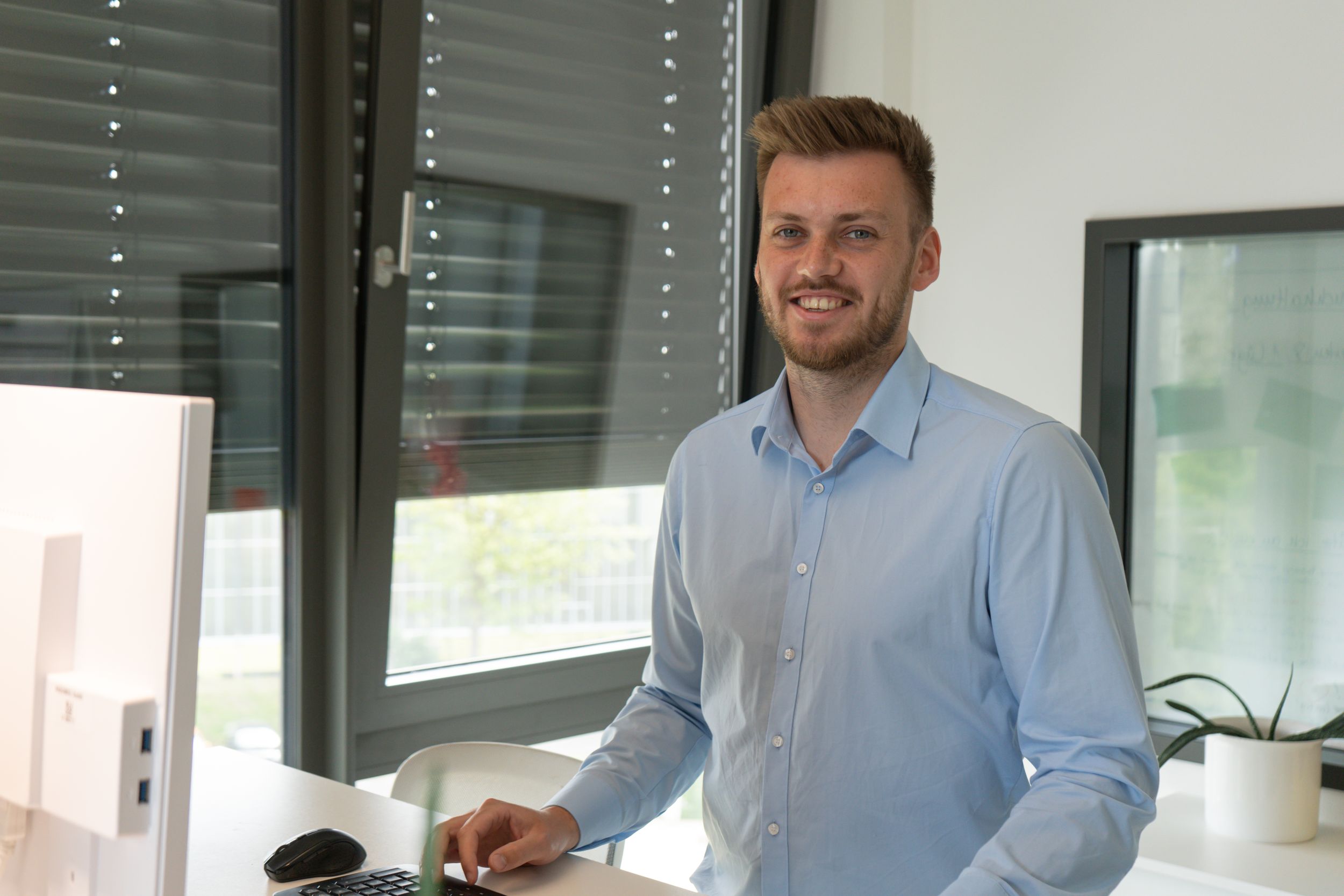 Junger Mann in hellblauem Hemd lächelt am Schreibtisch mit Computer und Tastatur im modernen Büro.