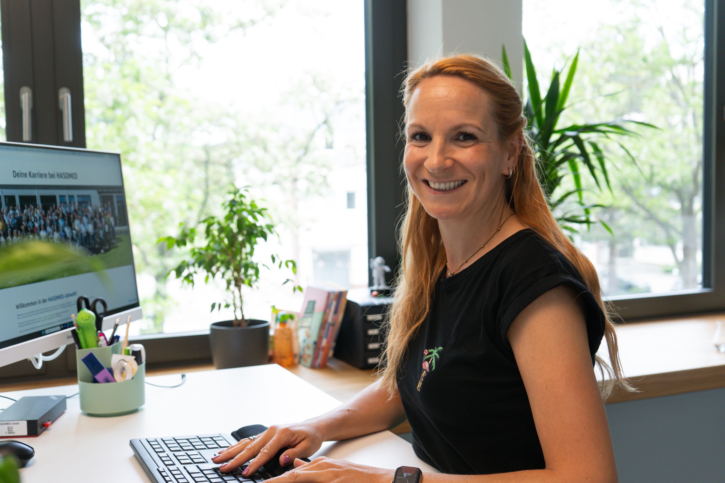 Lächelnde Frau sitzt mit den Händen auf der Tastatur an ihrem Schreibtisch vor einem Computer in einem hellen Büro mit großen Fenstern und grünen Pflanzen.