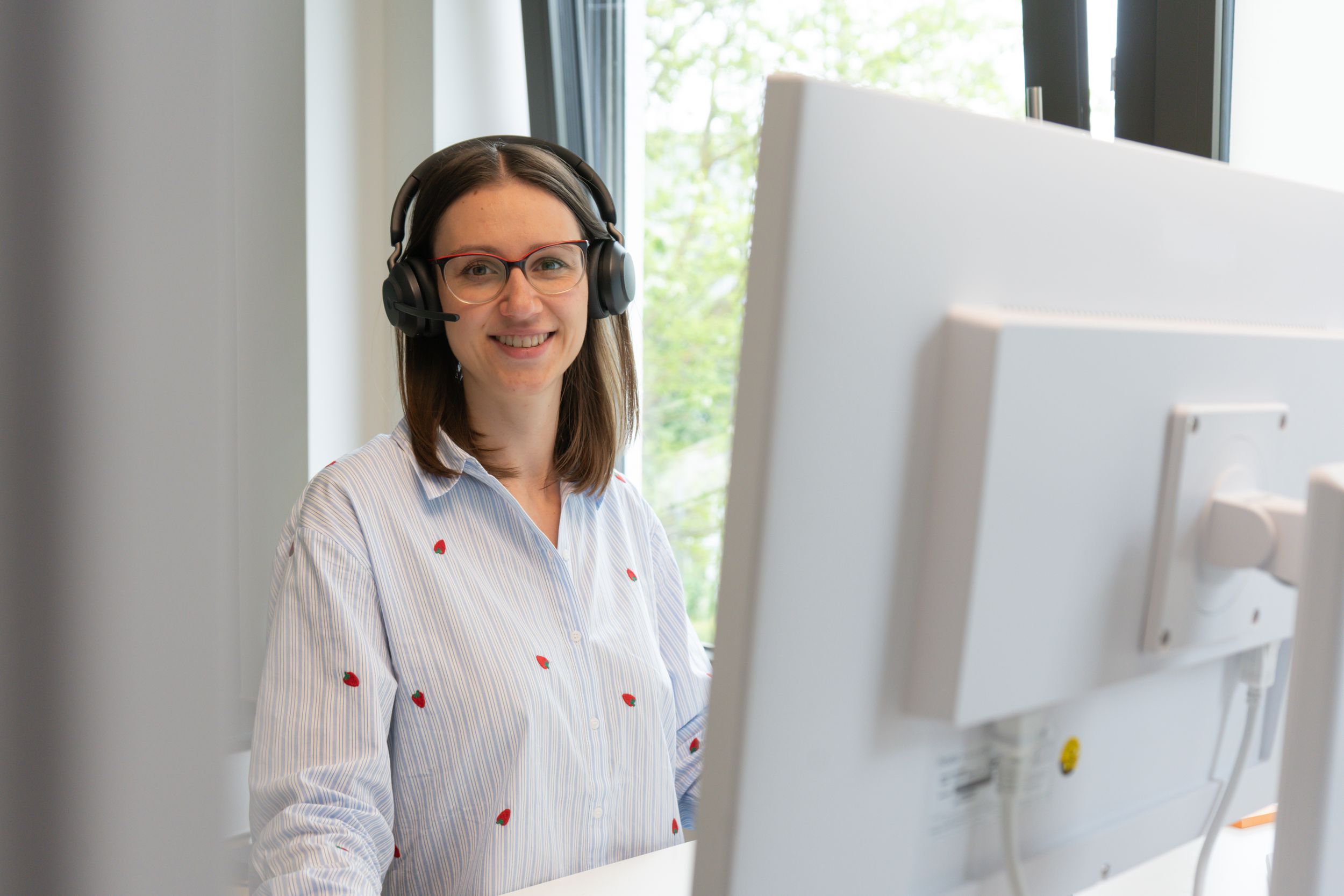 Lächelnde Frau mit Kopfhörer und Brille sitzt an einem Computer in einem hellen Büro mit Fensterblick auf grüne Bäume.