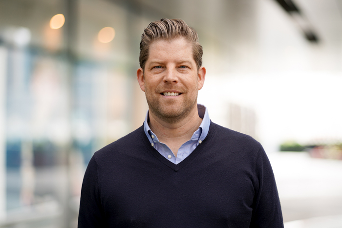 Porträt eines lächelnden Mannes mit hellbraunem Haar, der einen hellblauen Hemdkragen unter einem dunkelblauen Pullover trägt.