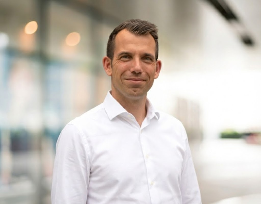 Lächelnder Mann mit braunen Haaren trägt ein weißes Hemd vor einem unscharfen Hintergrund.