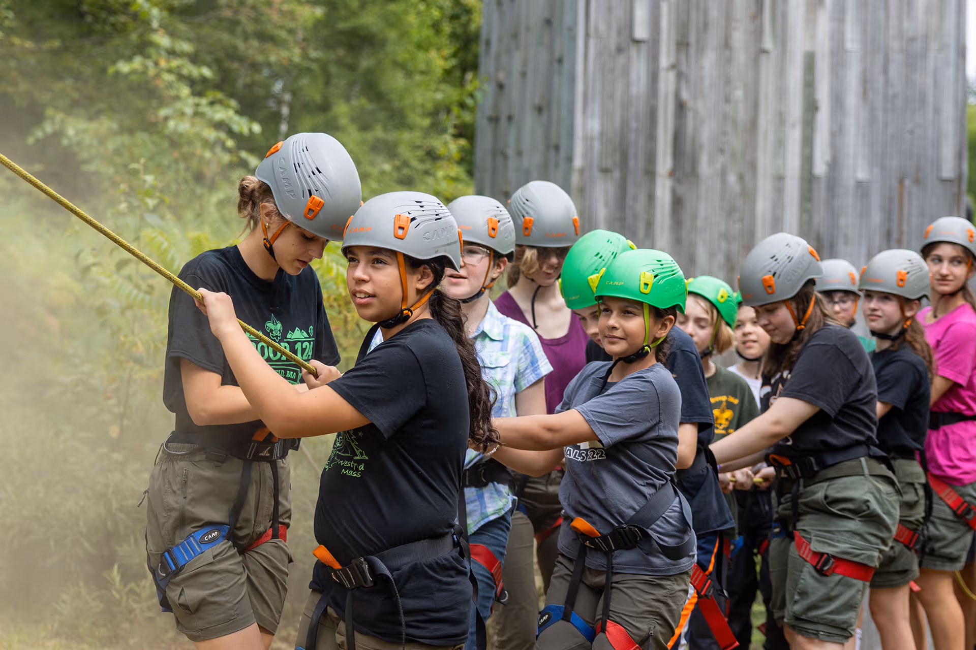 Group of children wearing helmets and harnesses participating in a rope climbing activity outdoors.