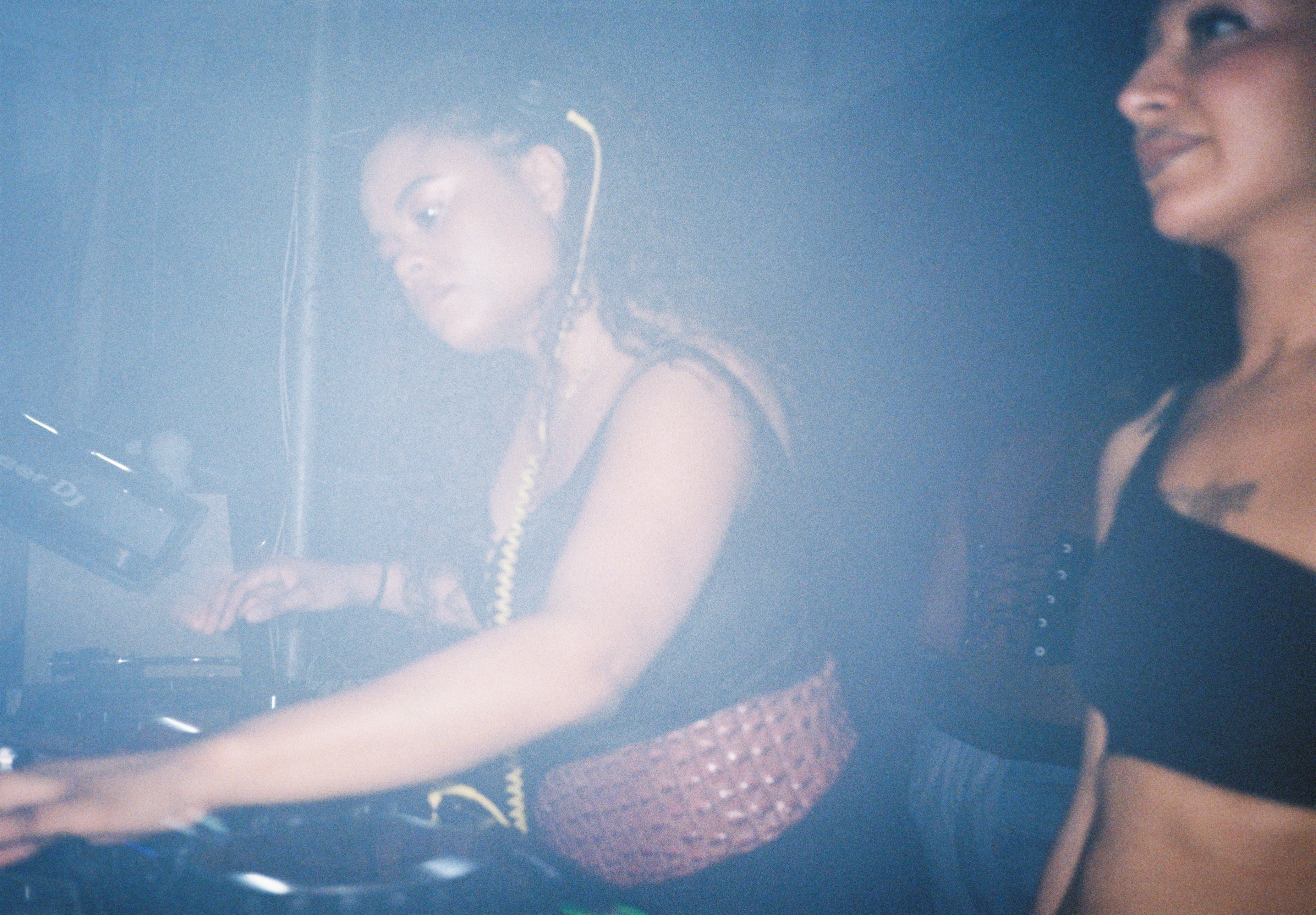 A female DJ with curly hair and tattoos adjusting headphones while a male DJ stands nearby in a dimly lit club.