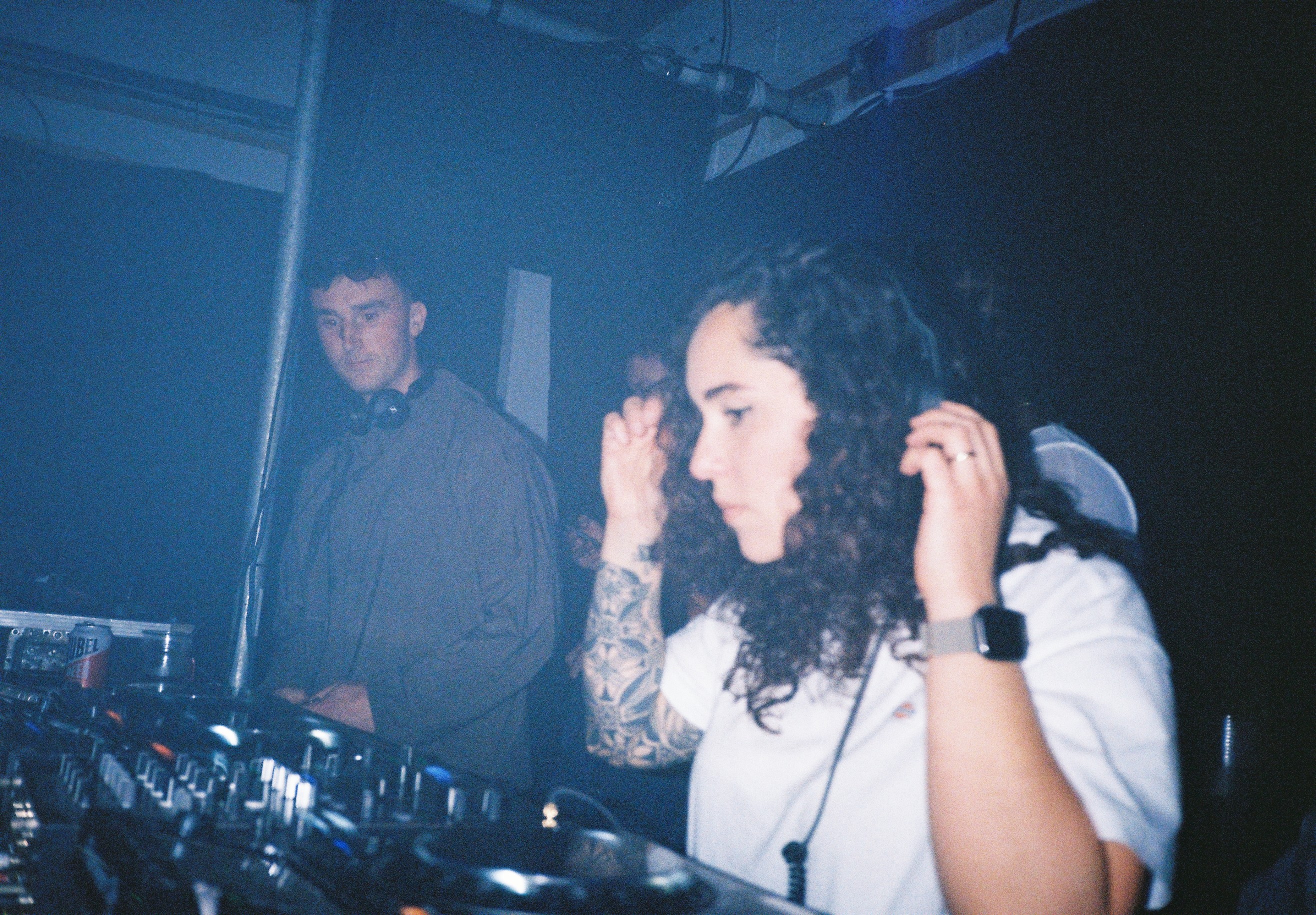A female DJ with curly hair and tattoos adjusting headphones while a male DJ stands nearby in a dimly lit club.