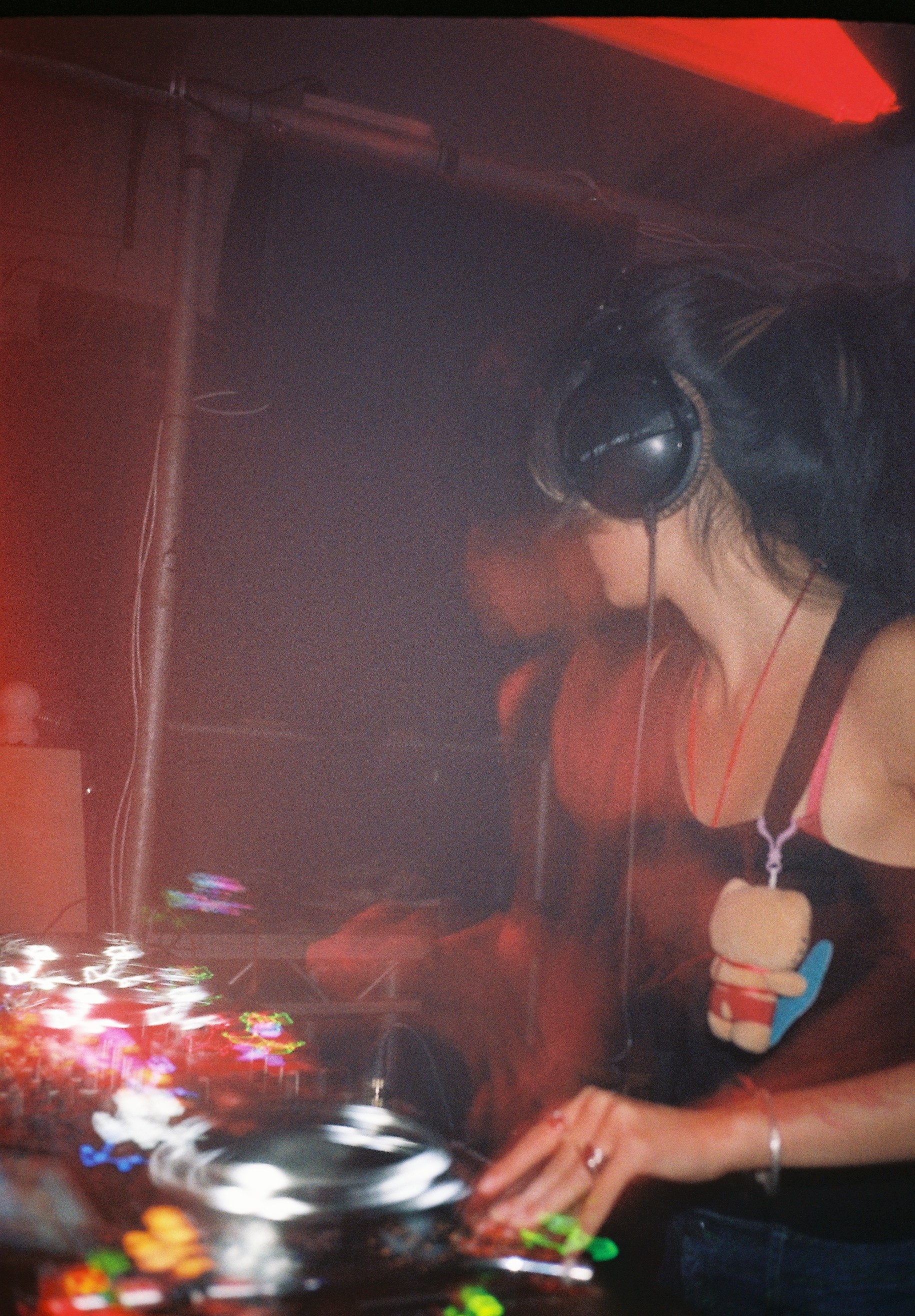 A female DJ with curly hair and tattoos adjusting headphones while a male DJ stands nearby in a dimly lit club.