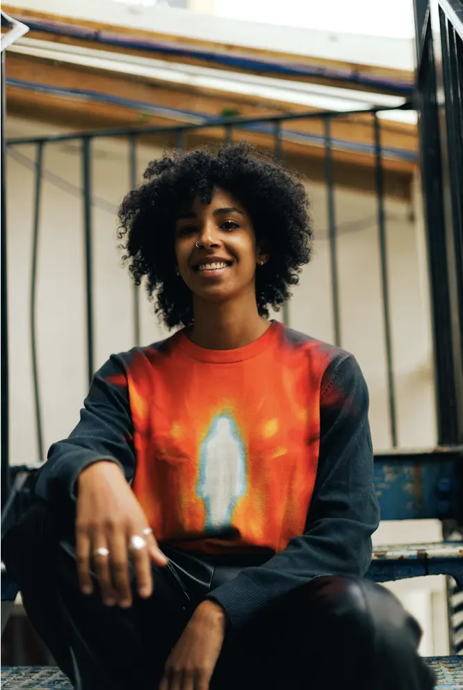 Person with curly hair smiles while seated on outdoor steps, wearing a long-sleeve sweatshirt with bold red and blue artwork on the front.