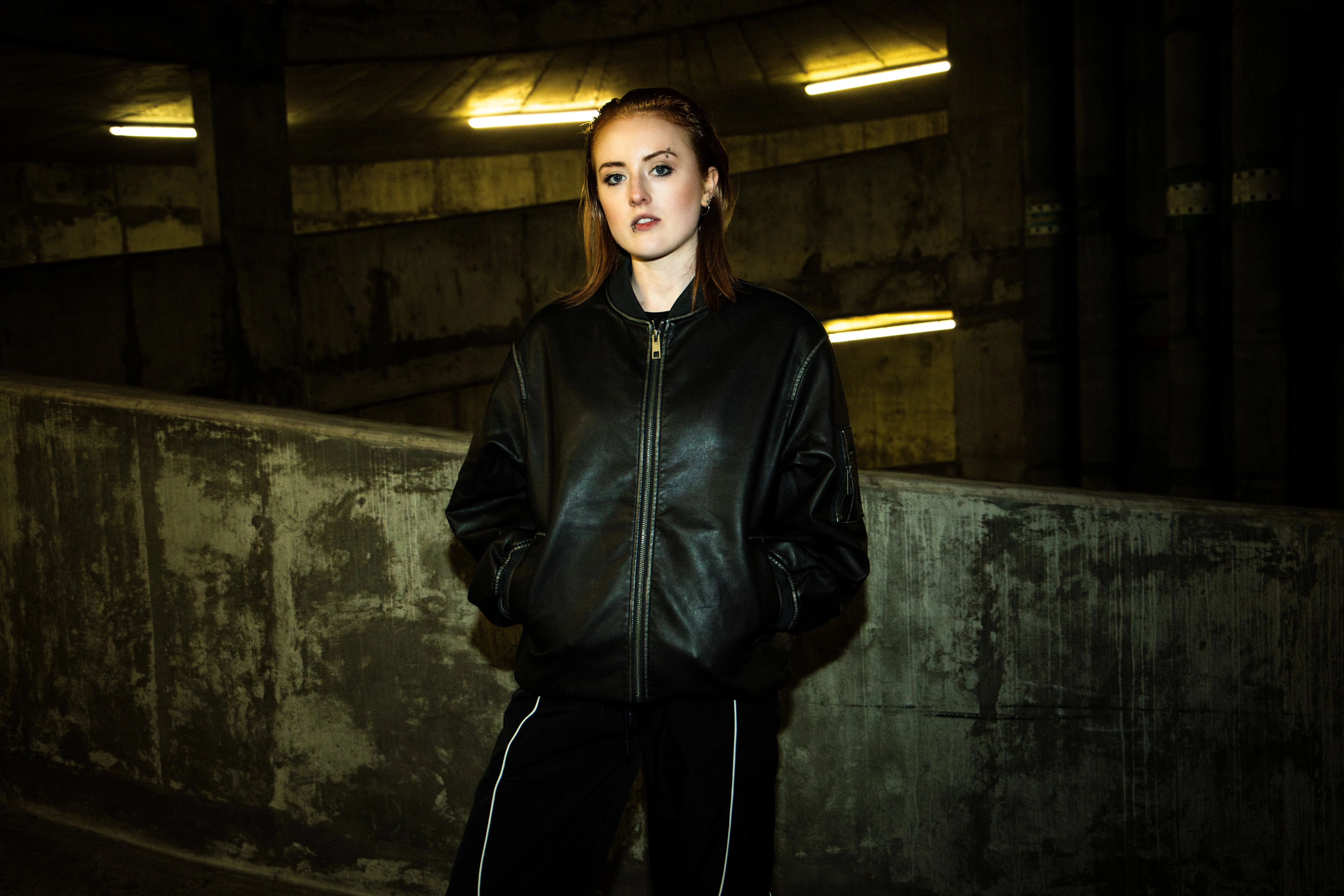 **Alt text:** > Person stands in a dimly lit concrete parking structure wearing a black leather jacket and dark trousers, hands in pockets under yellow overhead lights.