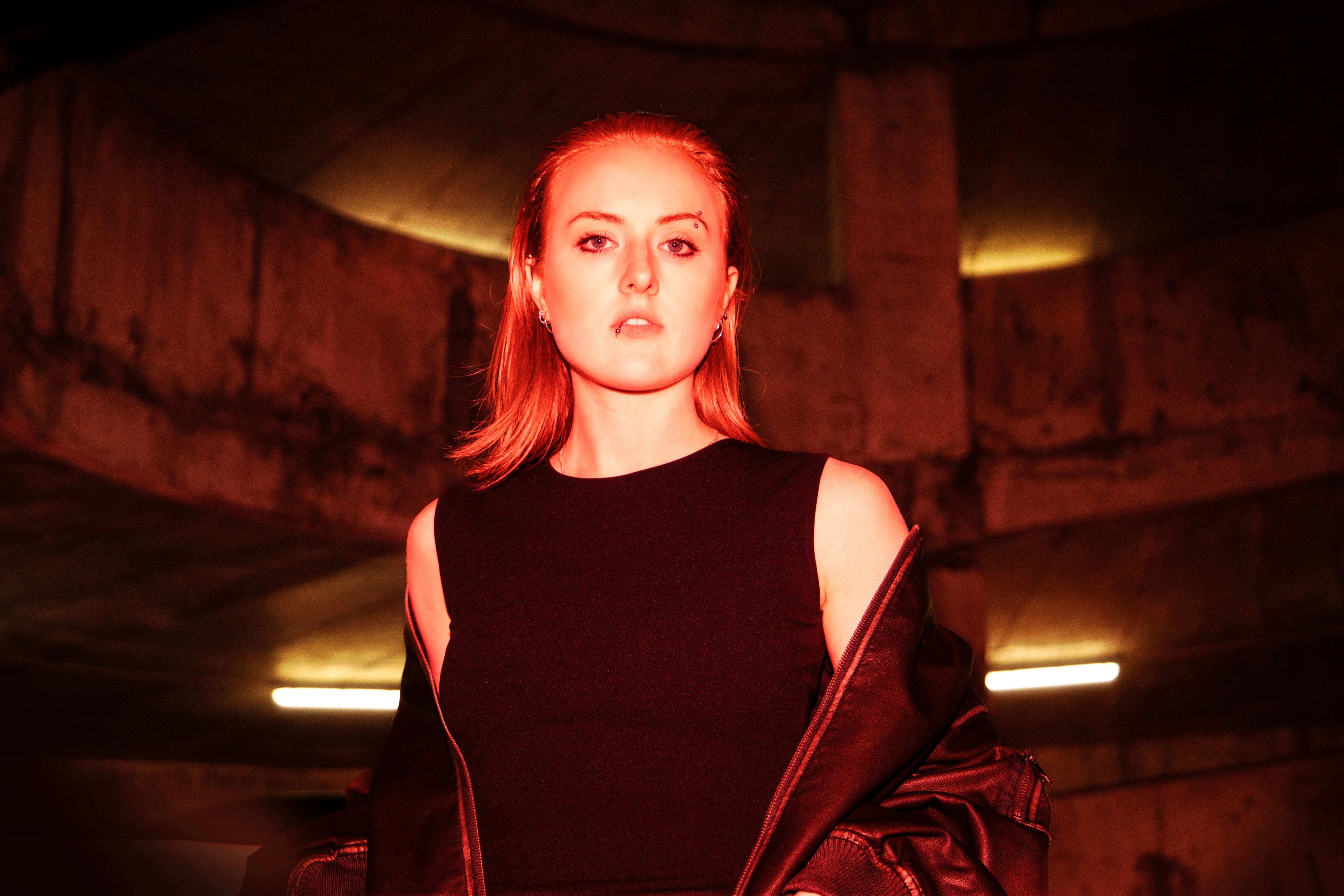**Alt text:** > Person stands in a dim concrete interior under red lighting, wearing a sleeveless black top with a jacket draped off their shoulders.