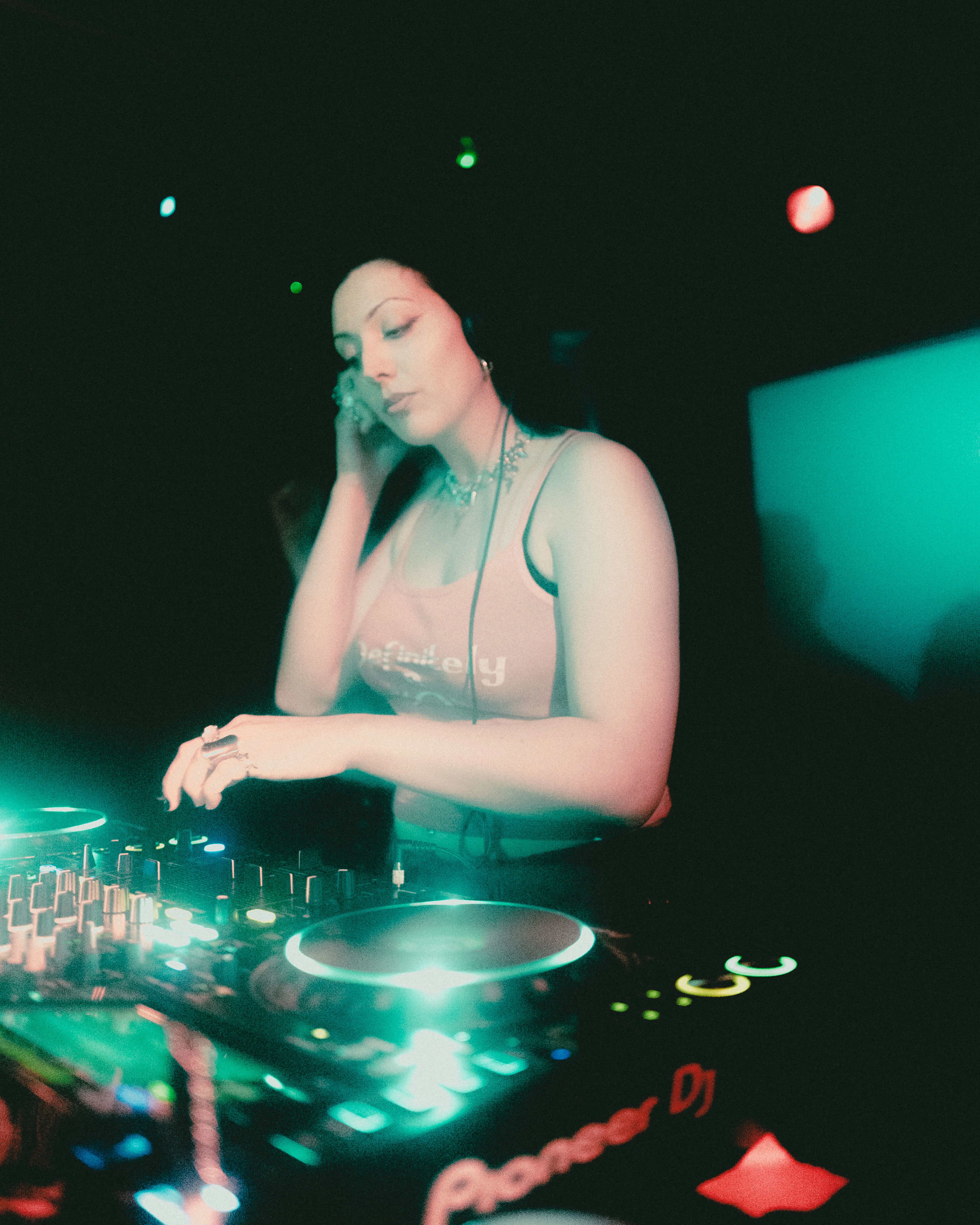 Person DJs in a dark club setting, wearing headphones and a sleeveless top while adjusting controls on a mixing deck under green and red stage lights.