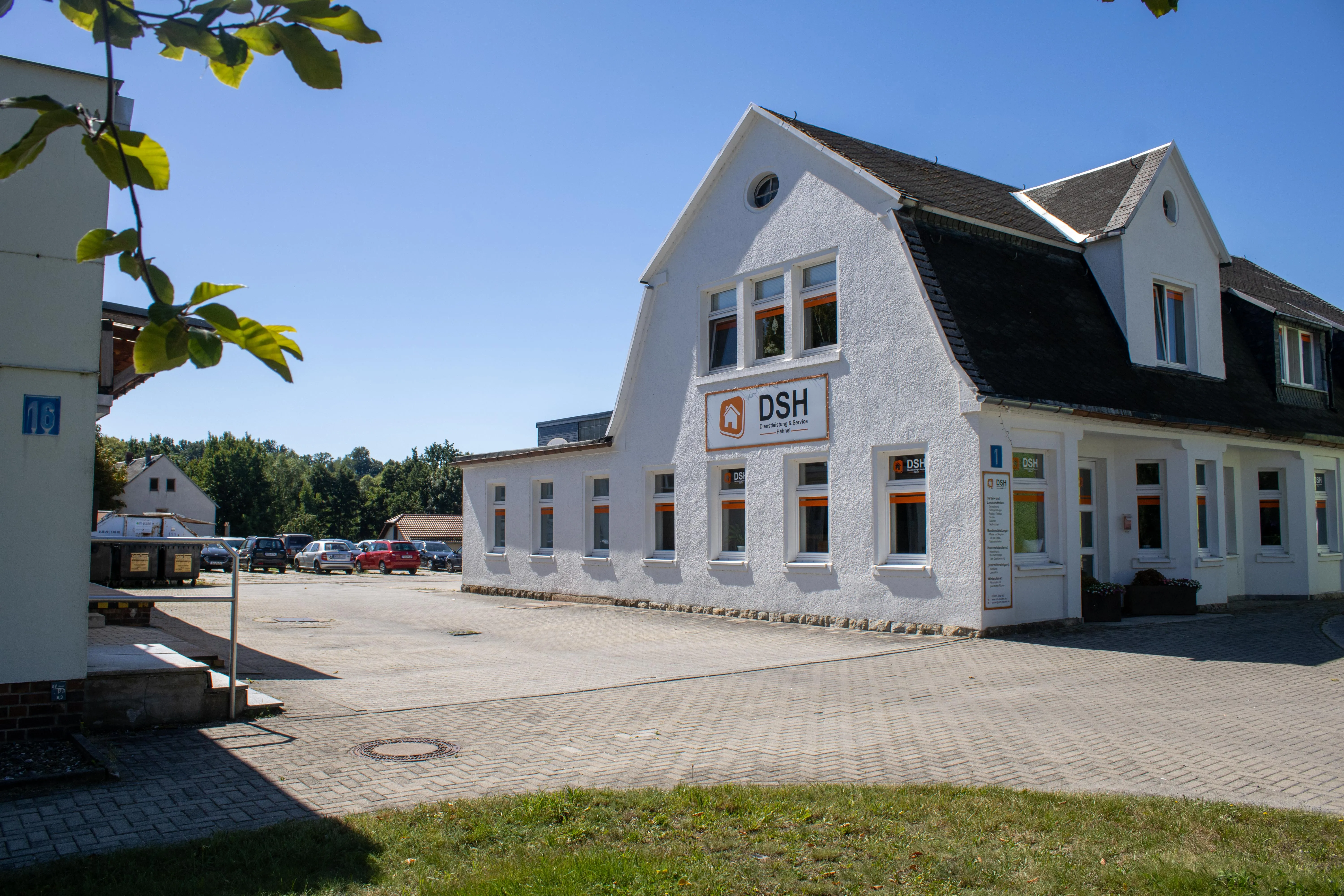 White building with DSH Dienstleistung & Service signage under a clear blue sky with parked cars and greenery in the background.