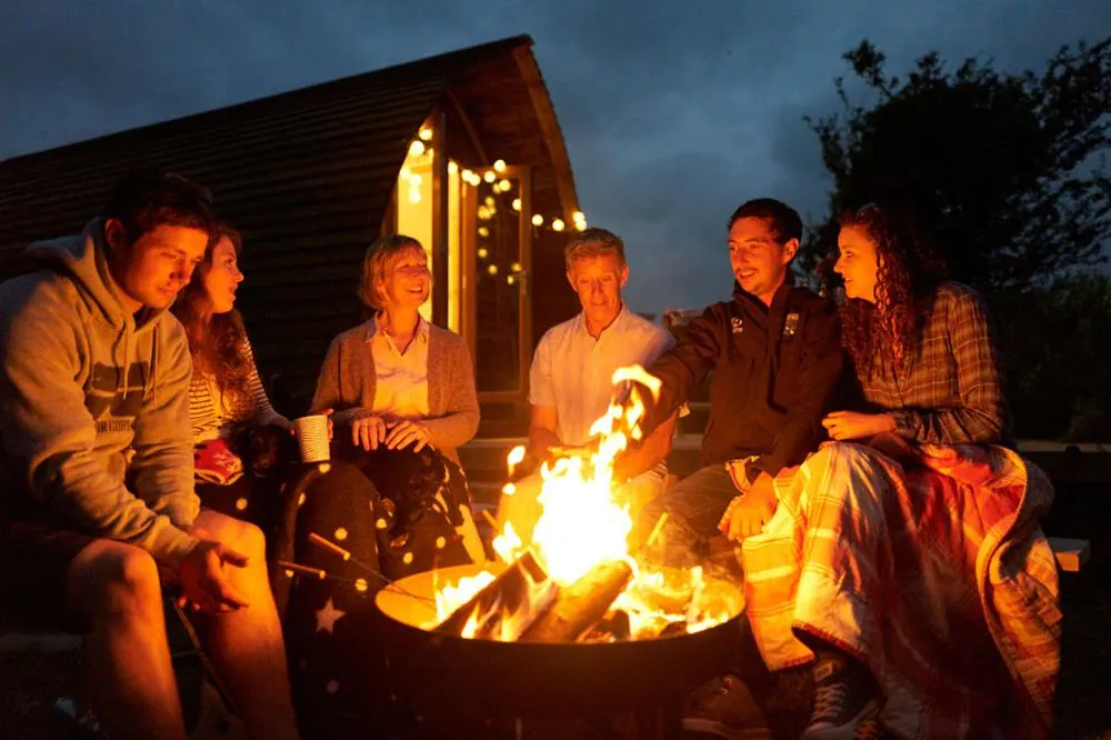 Friends gathered around a fire pit outside a Wigwam Holidays cabin at dusk
