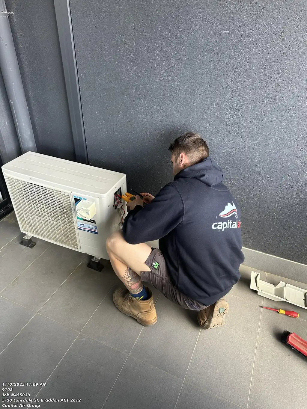 A technician wearing a Capital Air hoodie kneeling on the floor while servicing an outdoor Daikin air conditioning unit, using a multimeter and tools beside him.