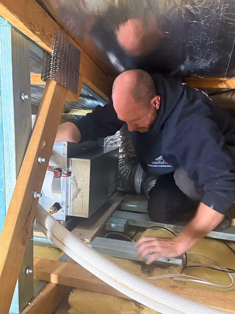Capital Air technician installing ducted heating and cooling equipment inside a roof space.